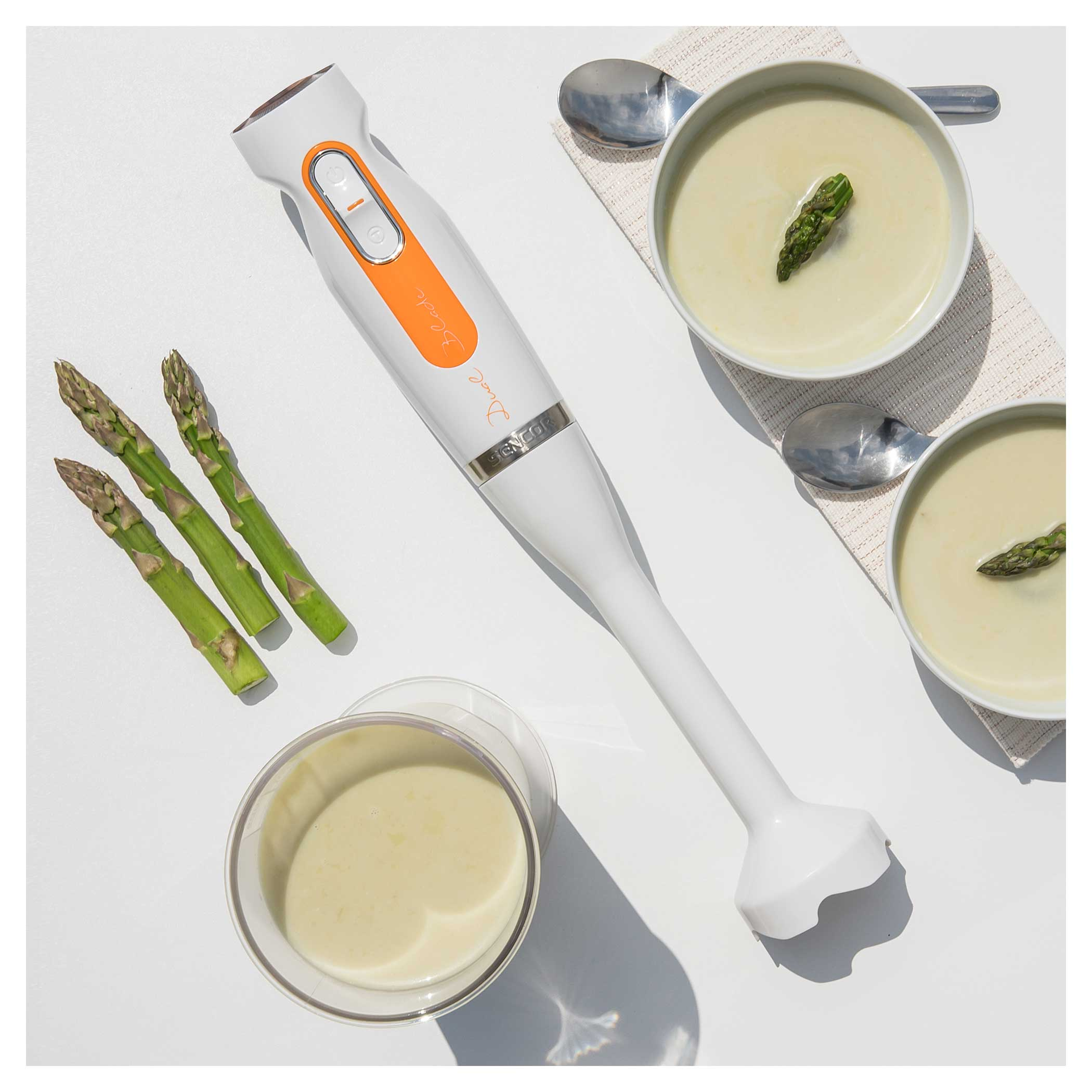 Frullatore bianco con dettagli arancioni vicino ad asparagi, ciotole di zuppa e un bicchiere.