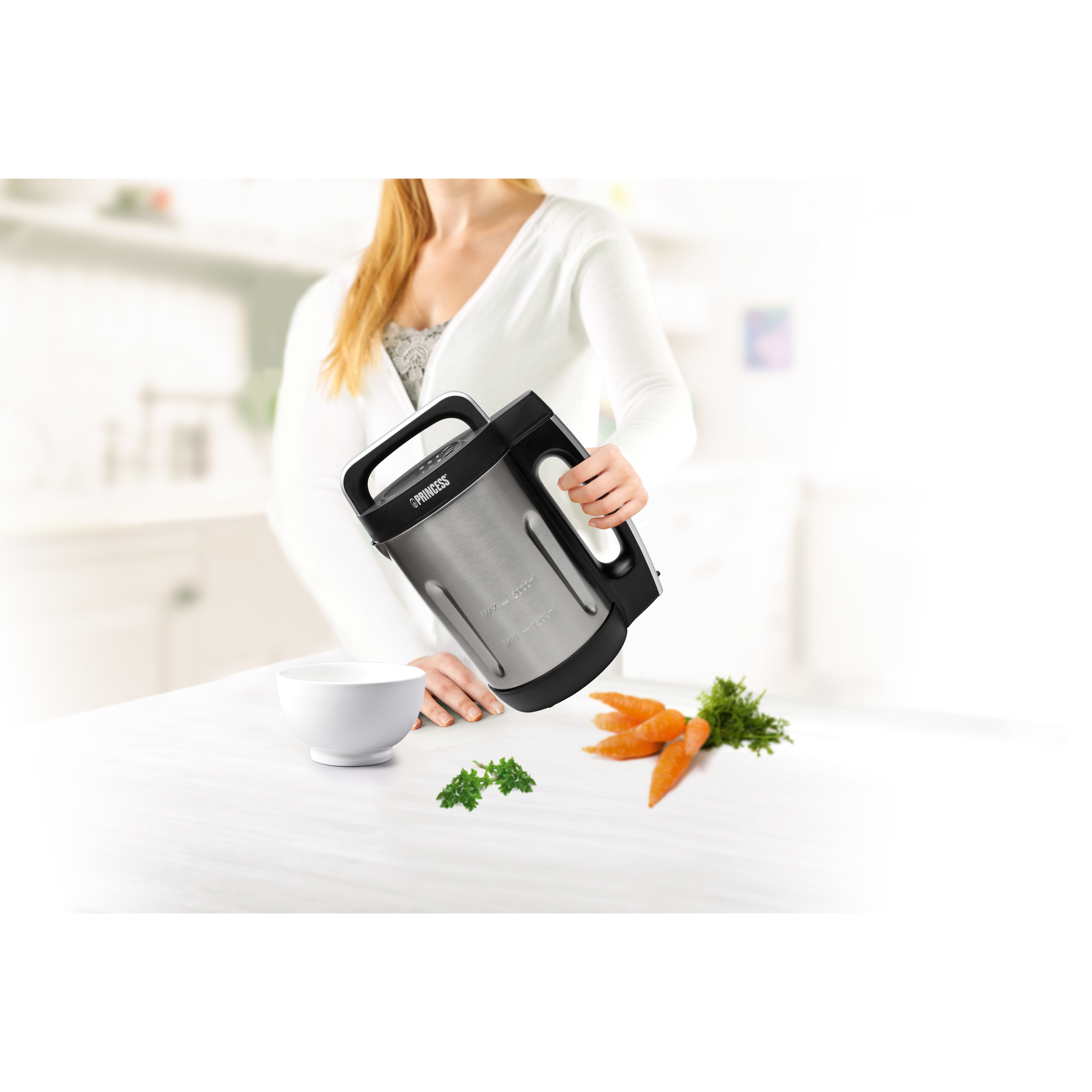 Una donna versa da un produttore di zuppa argento e nero in una ciotola bianca. Carote e prezzemolo sono accanto.