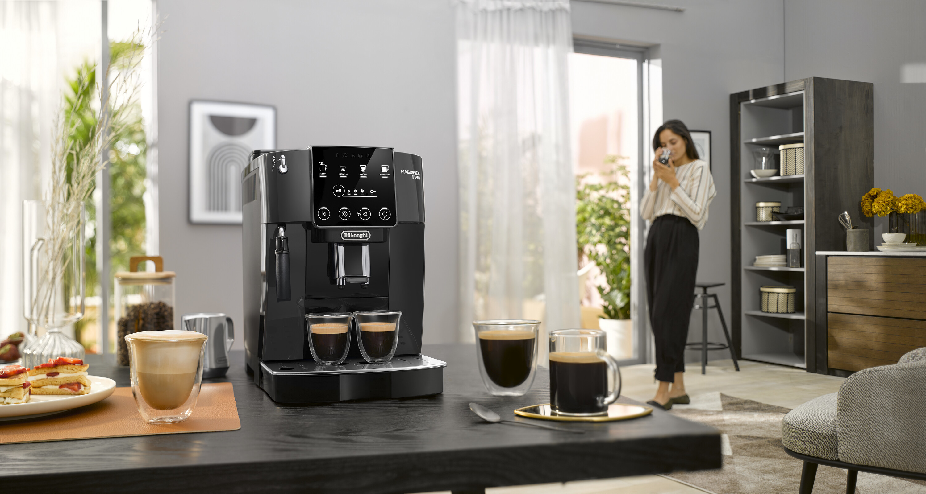 Macchina per caffè espresso nera sul tavolo, due tazze di caffè, donna sullo sfondo.