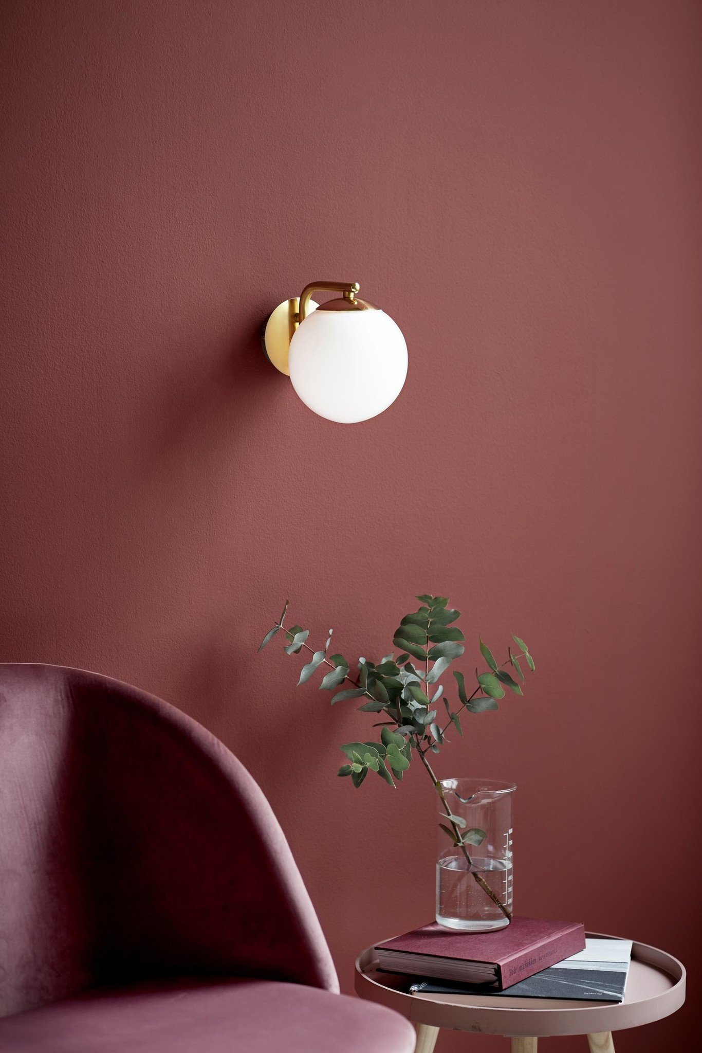Wandlampe mit weißem Globusschatten, grüne Pflanze und ein Buch auf einem Tisch vor einer weinroten Wand.