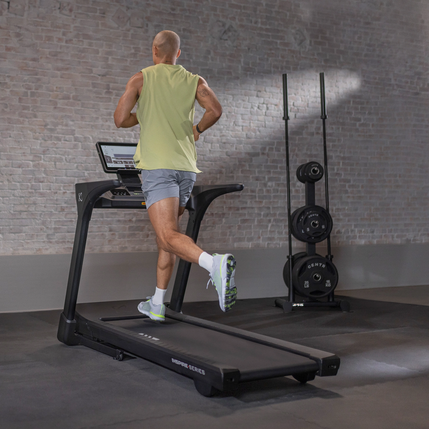 Ein Mann läuft in einem Fitnessstudio auf einem Laufband. Hinter ihm befinden sich Hantelständer und eine Ziegelmauer.