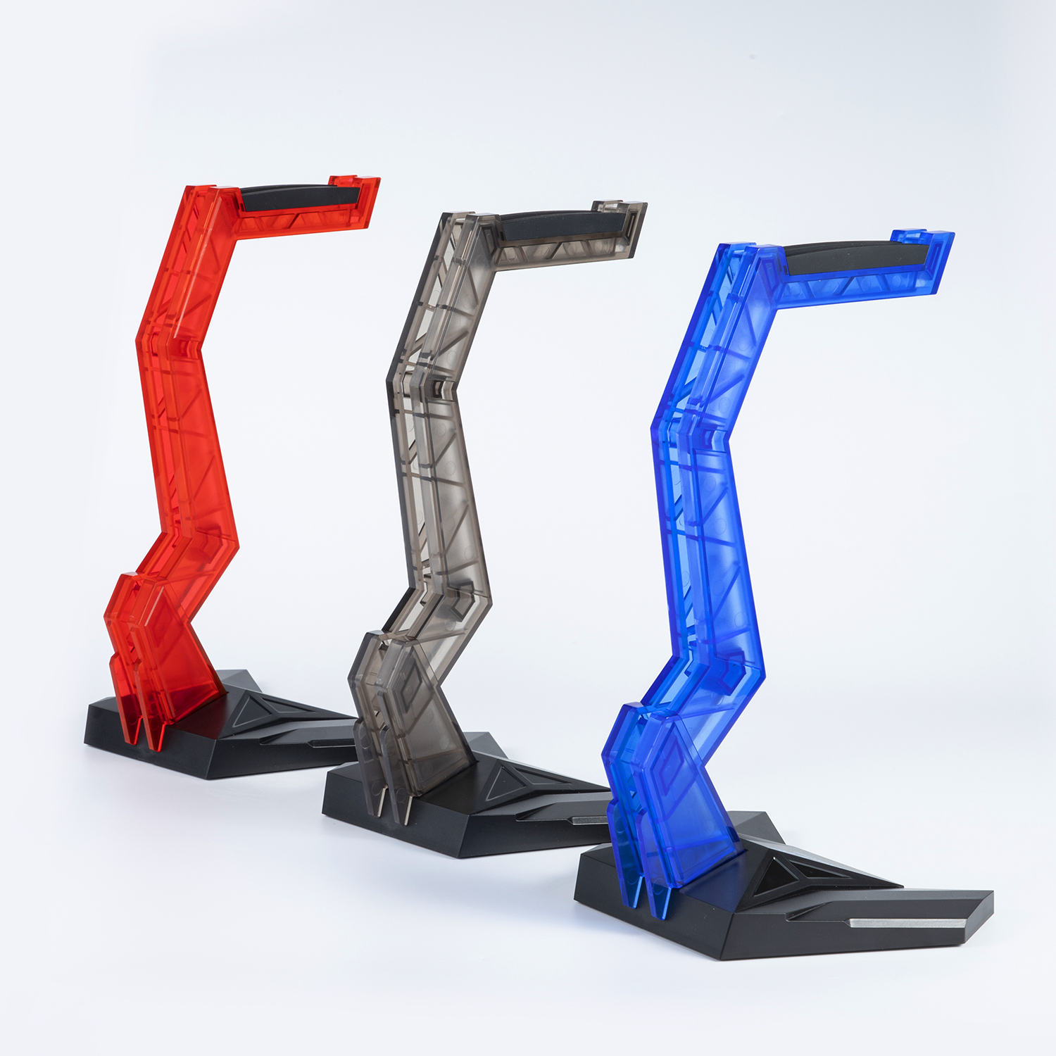 Tres soportes para auriculares gaming, rojo, gris y azul, sobre una superficie blanca.