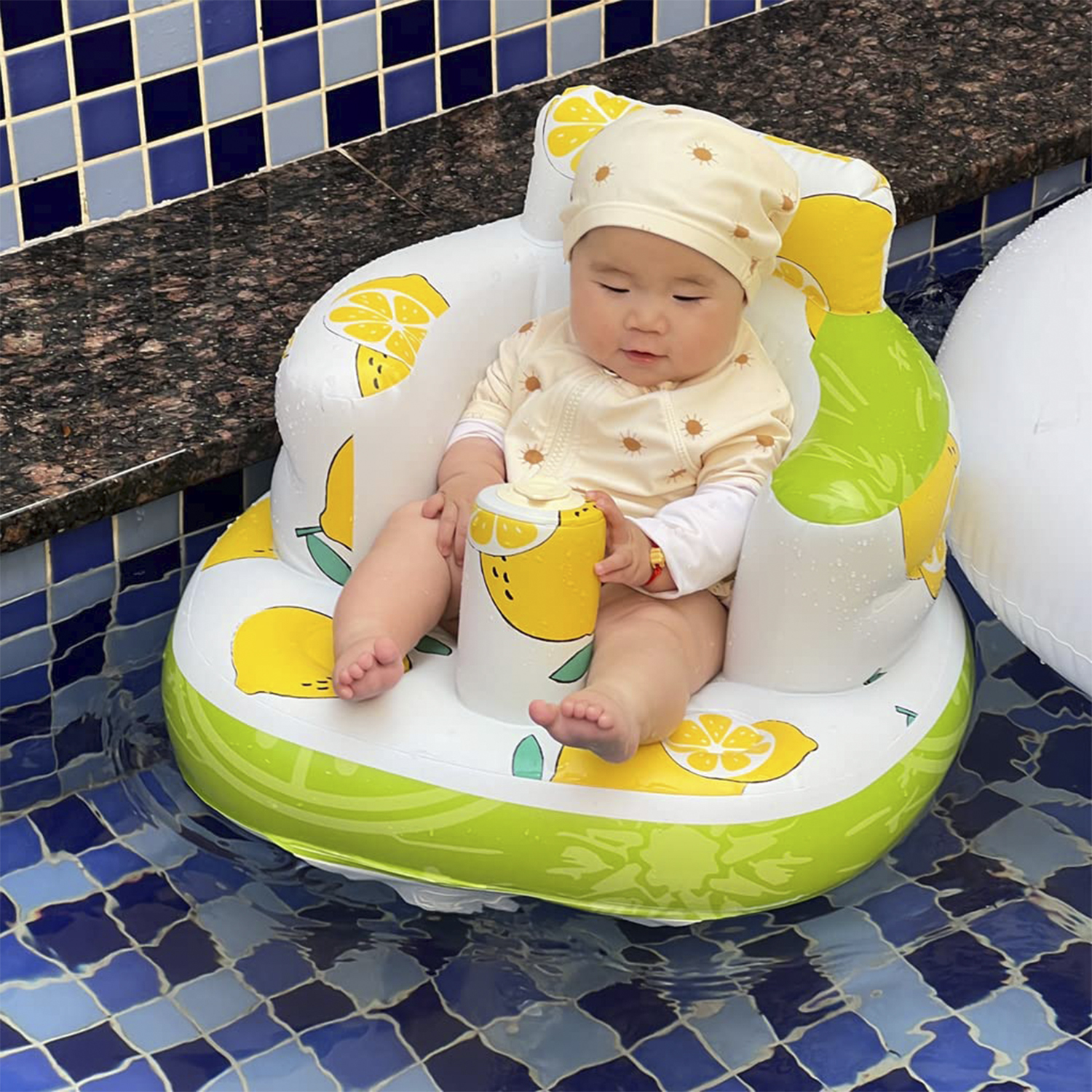 Bebé en una silla inflable con tema de limón en una piscina. El bebé lleva un traje de sol y un sombrero.