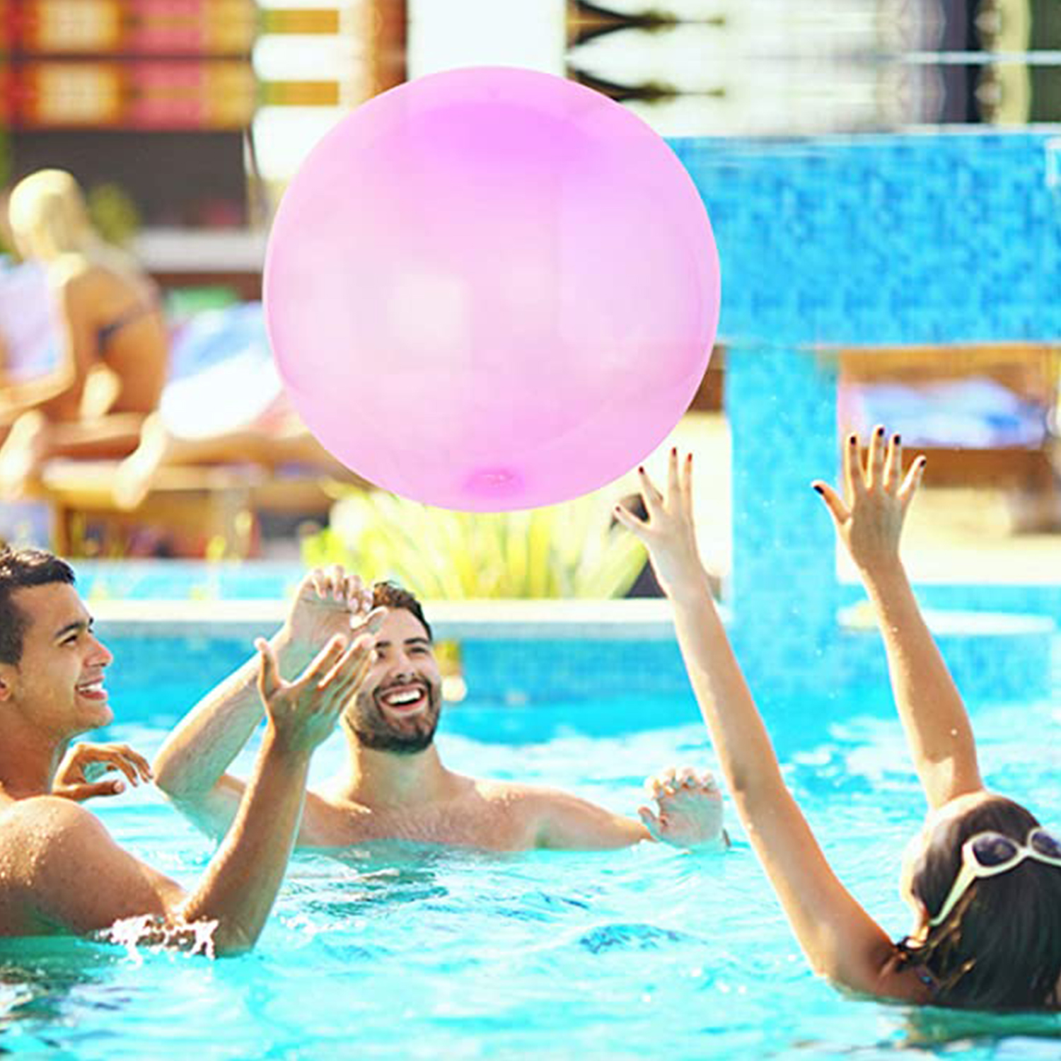 Tres personas en una piscina jugando con una gran pelota inflable rosa, día soleado.