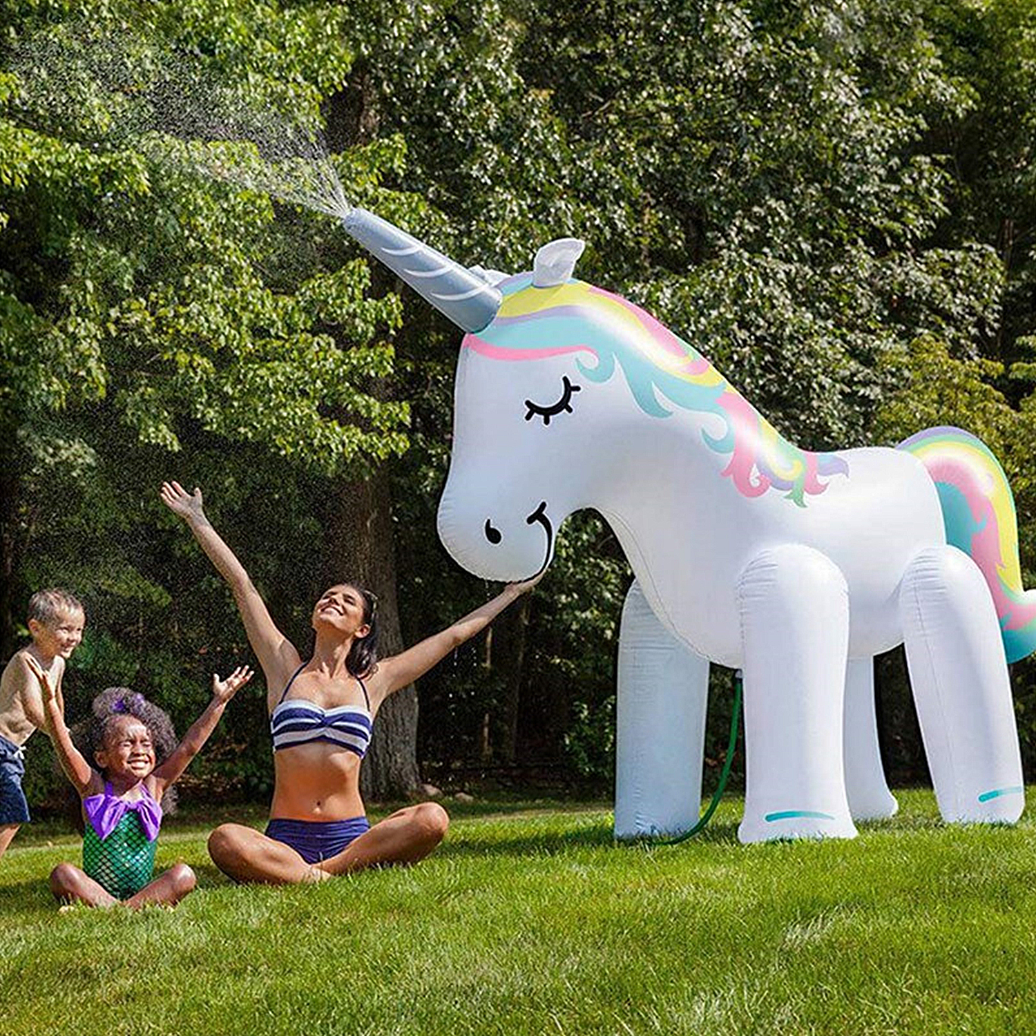 Un gran unicornio inflable rociando agua con una mujer y niños en un jardín.