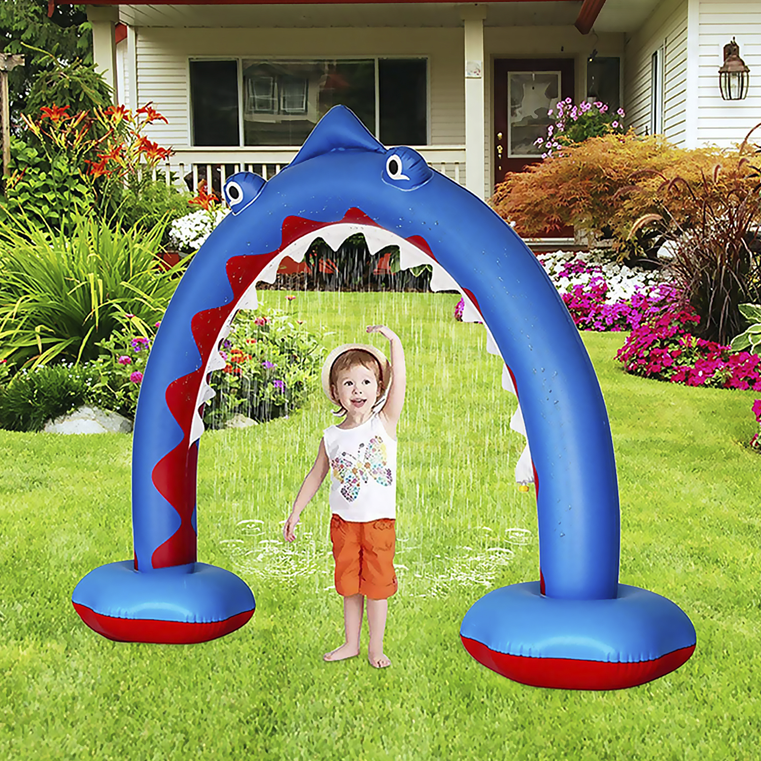 Un niño se para debajo de un rociador de agua inflable con forma de tiburón en un césped.