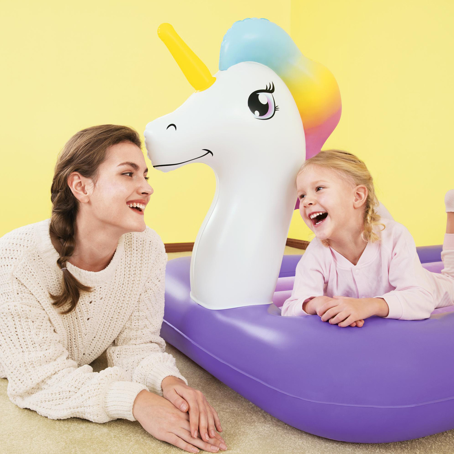 Una donna e una bambina sono sdraiate vicino a un giocattolo gonfiabile a forma di unicorno, entrambe sorridenti.