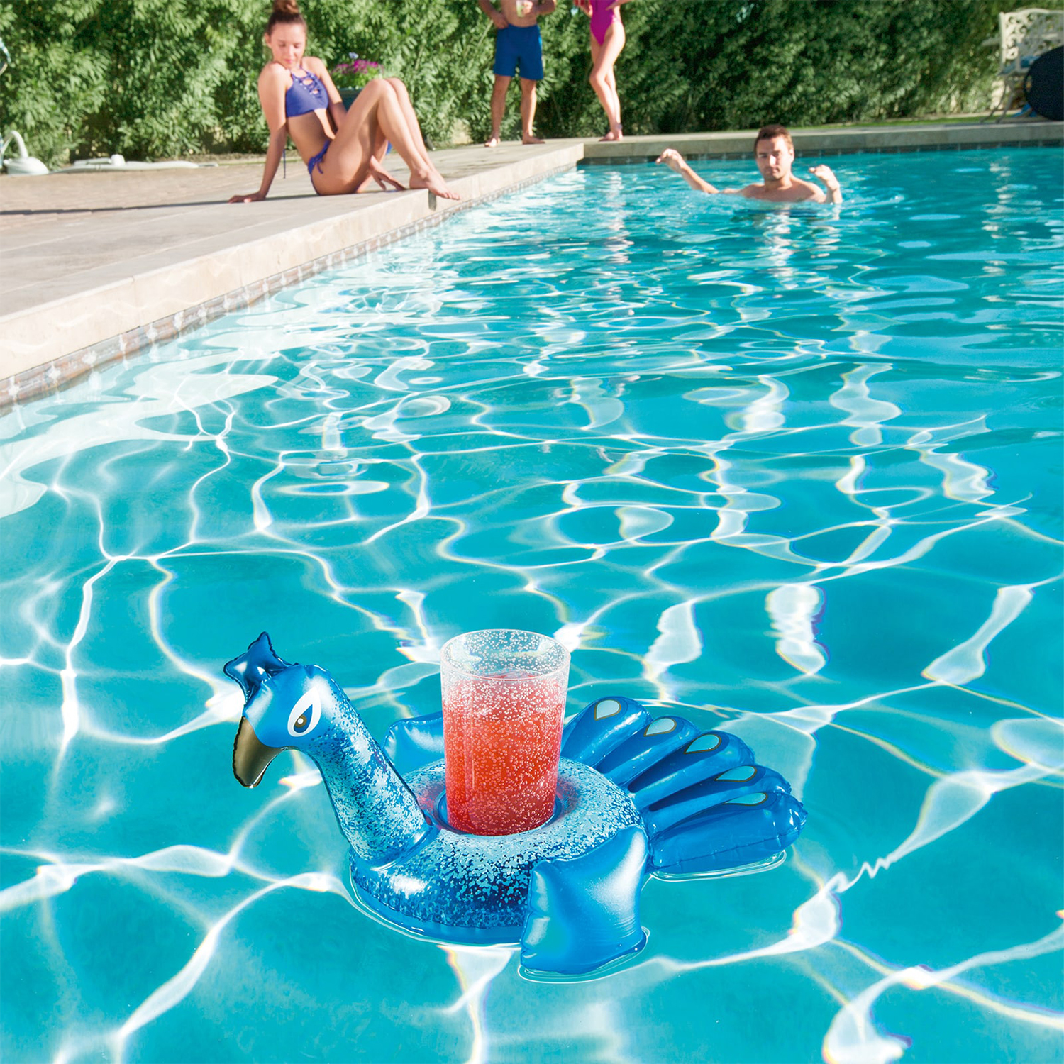 Una piscina con un galleggiante a forma di pavone blu con una bevanda. Persone vicino alla piscina, che si rilassano.
