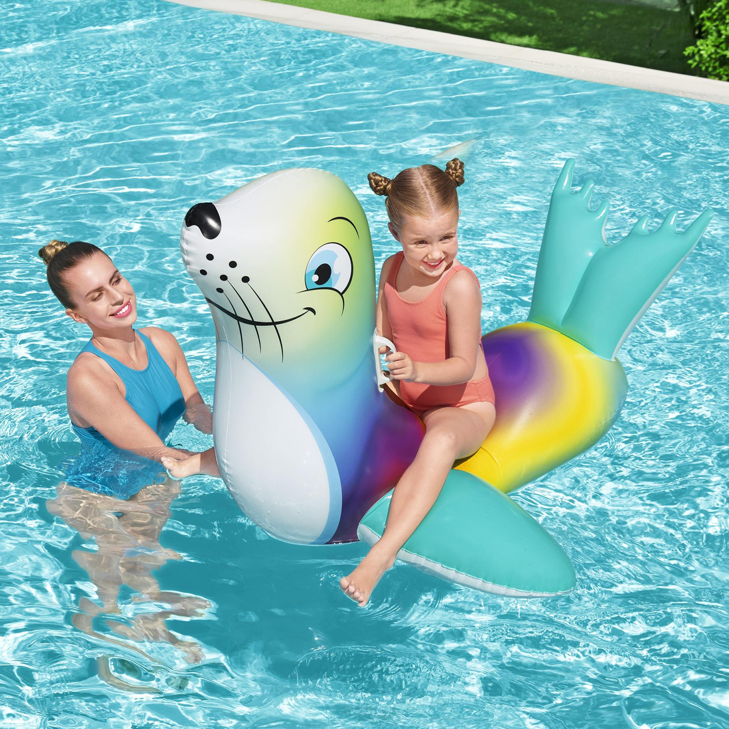 Una donna e una bambina cavalcano una foca gonfiabile in piscina. Acqua blu limpida.