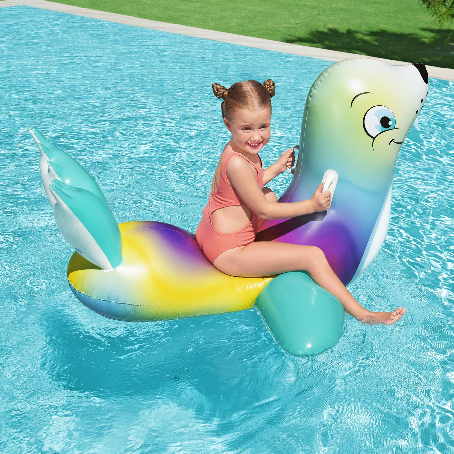 Una ragazza cavalca una foca gonfiabile in una piscina. La foca è colorata e la ragazza sorride.