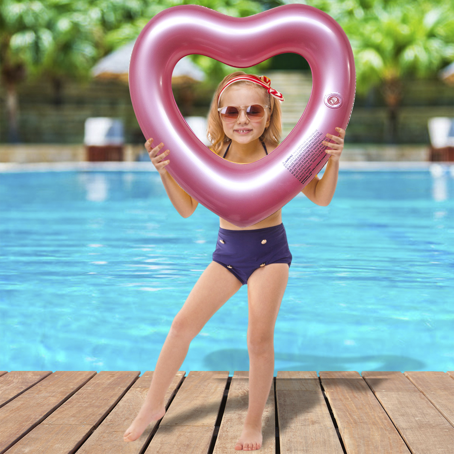 Niña con gafas de sol y traje de baño sosteniendo un flotador rosa en forma de corazón en la piscina.