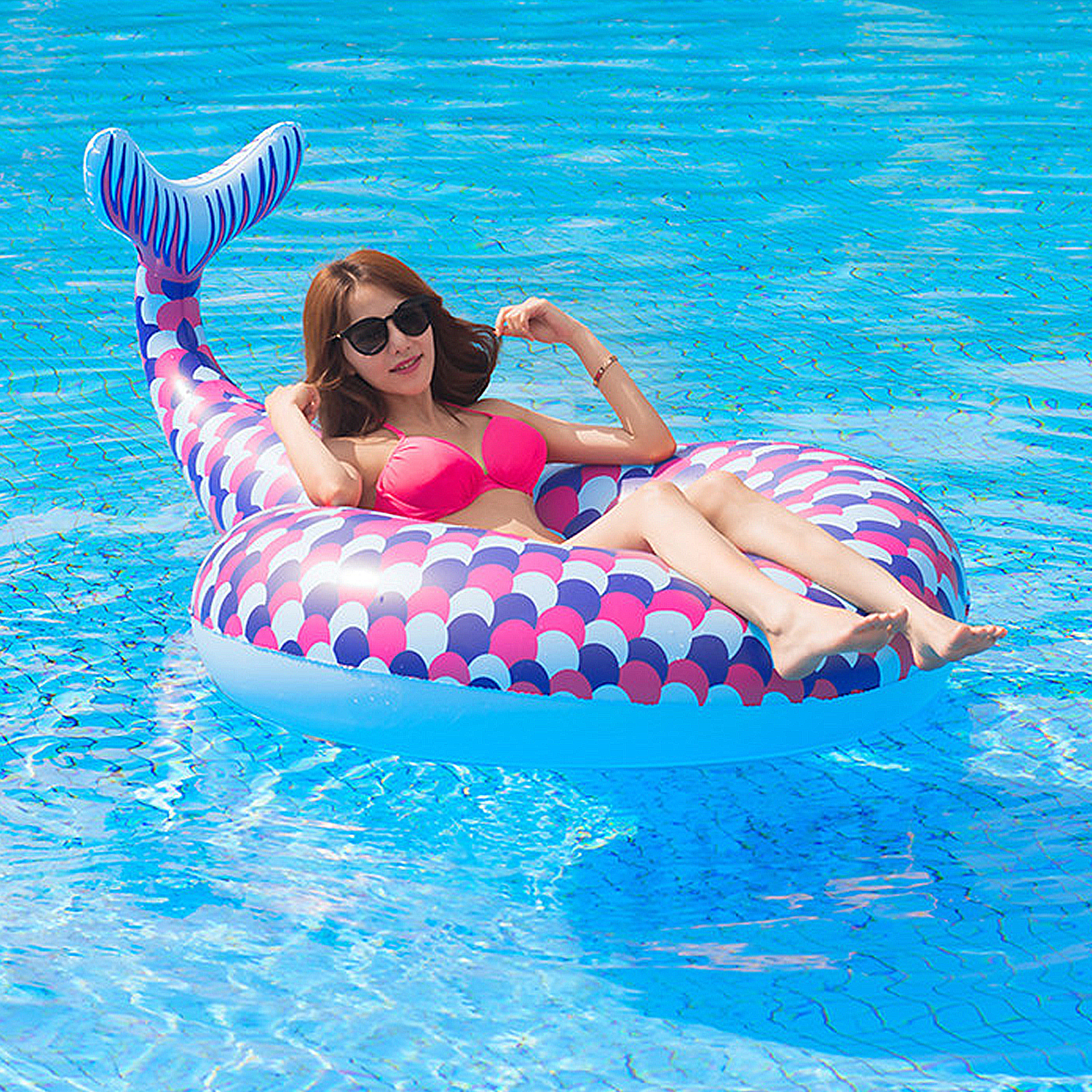 Una donna in bikini rosa su un galleggiante a sirena in piscina. Indossa occhiali da sole e sorride. Cielo blu.
