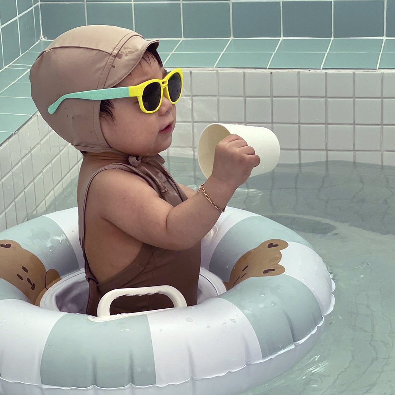 Bebé en un flotador, con gafas de sol y sombrero, sosteniendo una taza en agua.