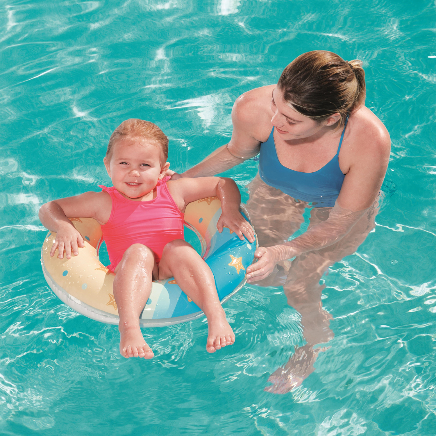 Una niña pequeña está sentada en un anillo inflable amarillo y azul en una piscina, con una mujer cerca.