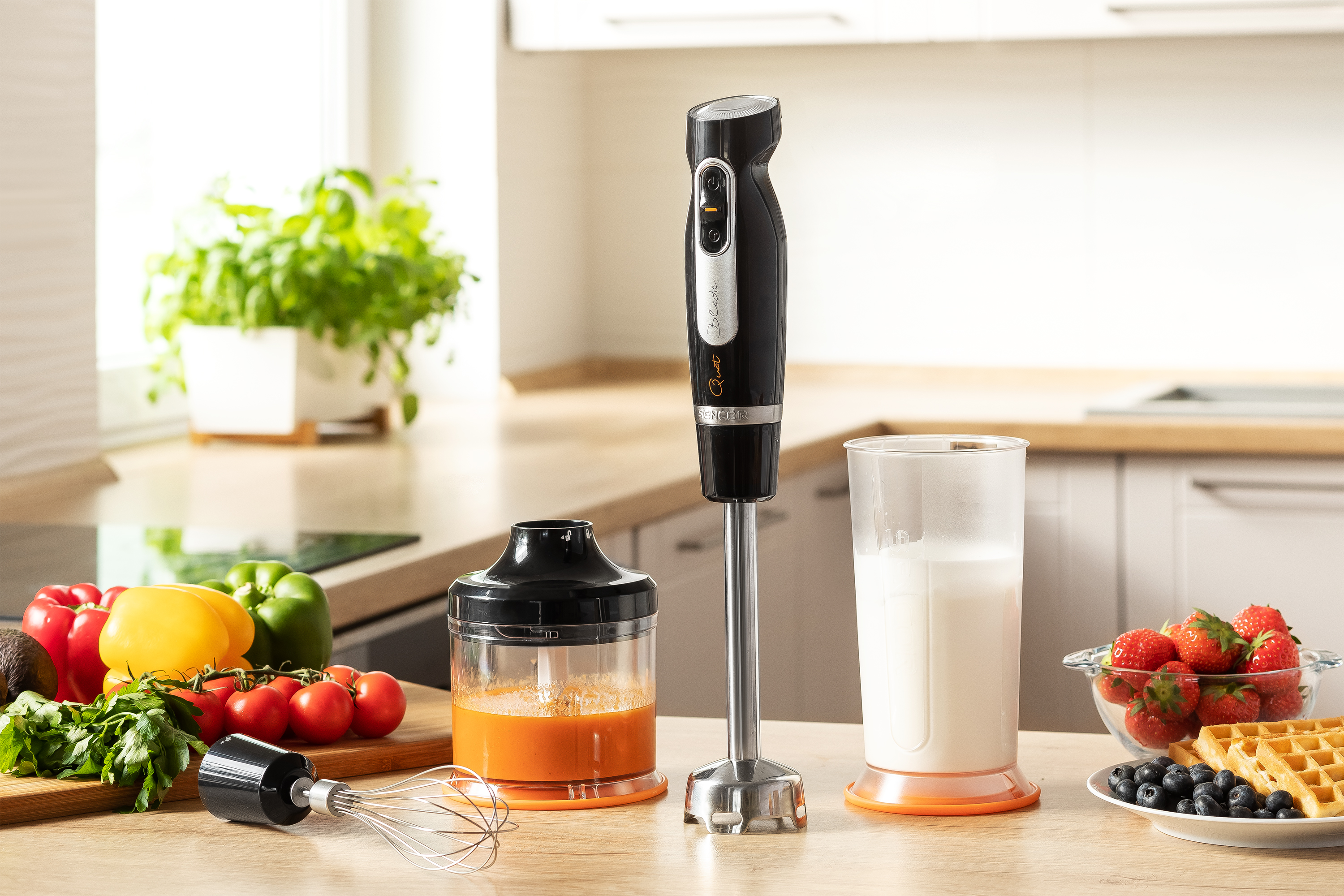 Frullatore a immersione con dettagli neri e arancioni, accanto a ingredienti e un bicchiere di latte.