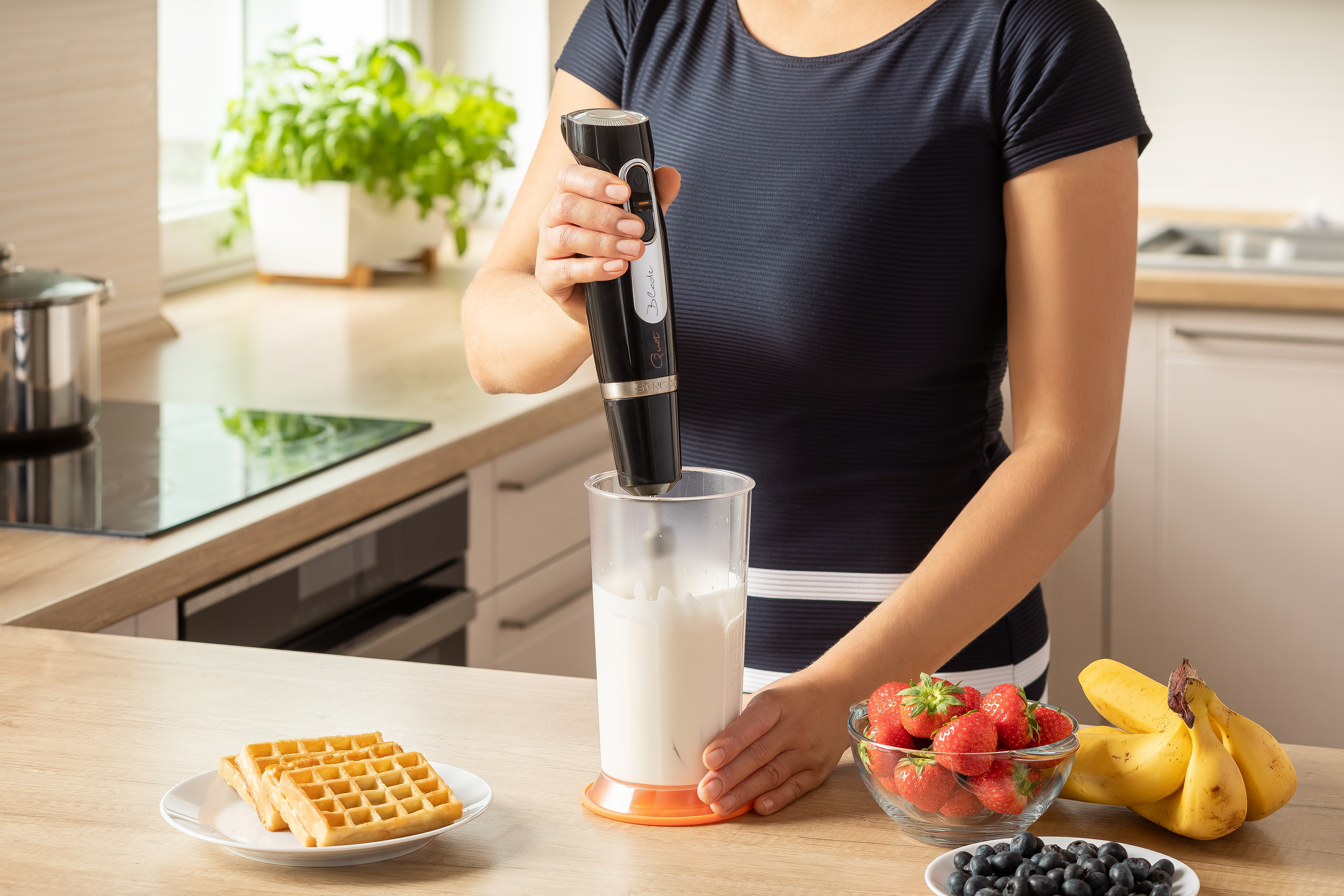 Osoba miksuje mleko w kubku za pomocą blendera ręcznego na blacie.