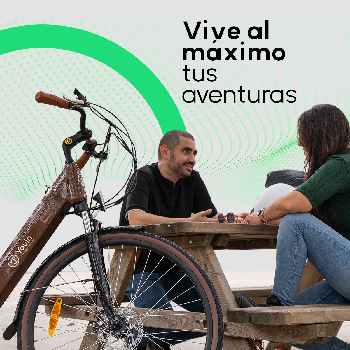 Dwie osoby siedzą przy stole piknikowym z rowerem. Tekst mówi: 'Vive al maximo tus aventuras'.