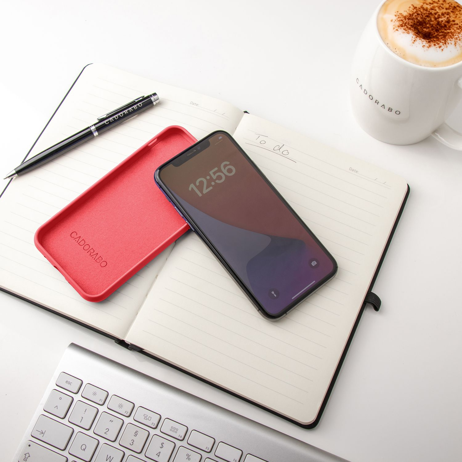 Ein Smartphone, eine rote Hülle, ein Stift und ein Becher auf einem Notizbuch, mit Tastatur im Hintergrund.