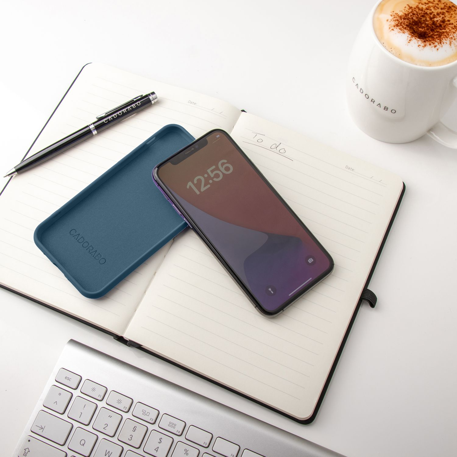 Notizbuch, Telefon, Stift, Becher und Tastatur auf einem weißen Schreibtisch, Draufsicht.
