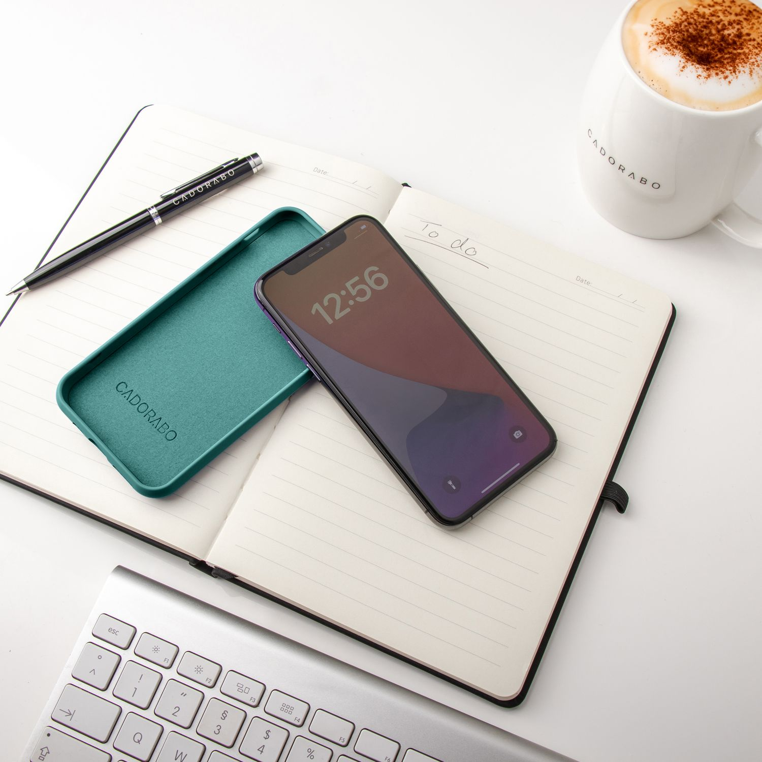 Ein Notizbuch, ein Telefon und eine Tasse mit Kaffee auf weißem Untergrund. Stift und Hülle sind ebenfalls vorhanden.