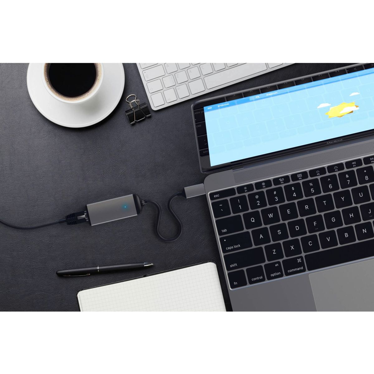Laptop collegato a un adattatore su una scrivania nera con penna e tazza di caffè.