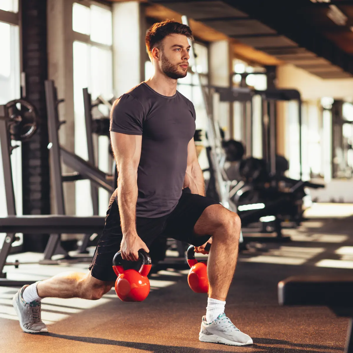 Ein Mann im Fitnessstudio macht Ausfallschritte mit roten Kettlebells. Er trägt Sportkleidung und schaut weg.