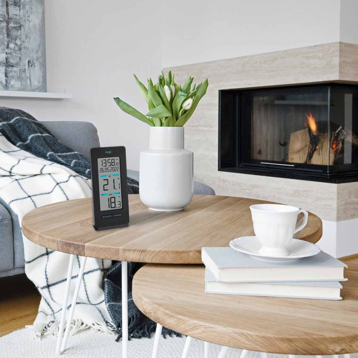 Ein digitales Thermometer auf einem Holztisch, neben einer weißen Vase mit Tulpen und einem Kamin.