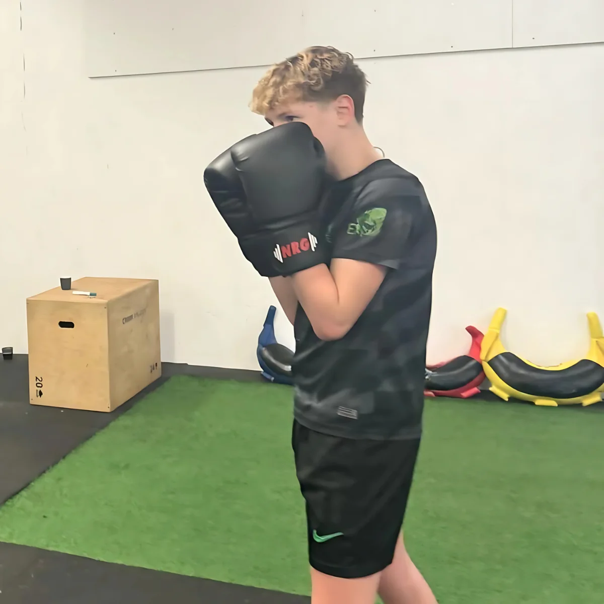 A young man wearing boxing gloves, covering his face in a gym setting.