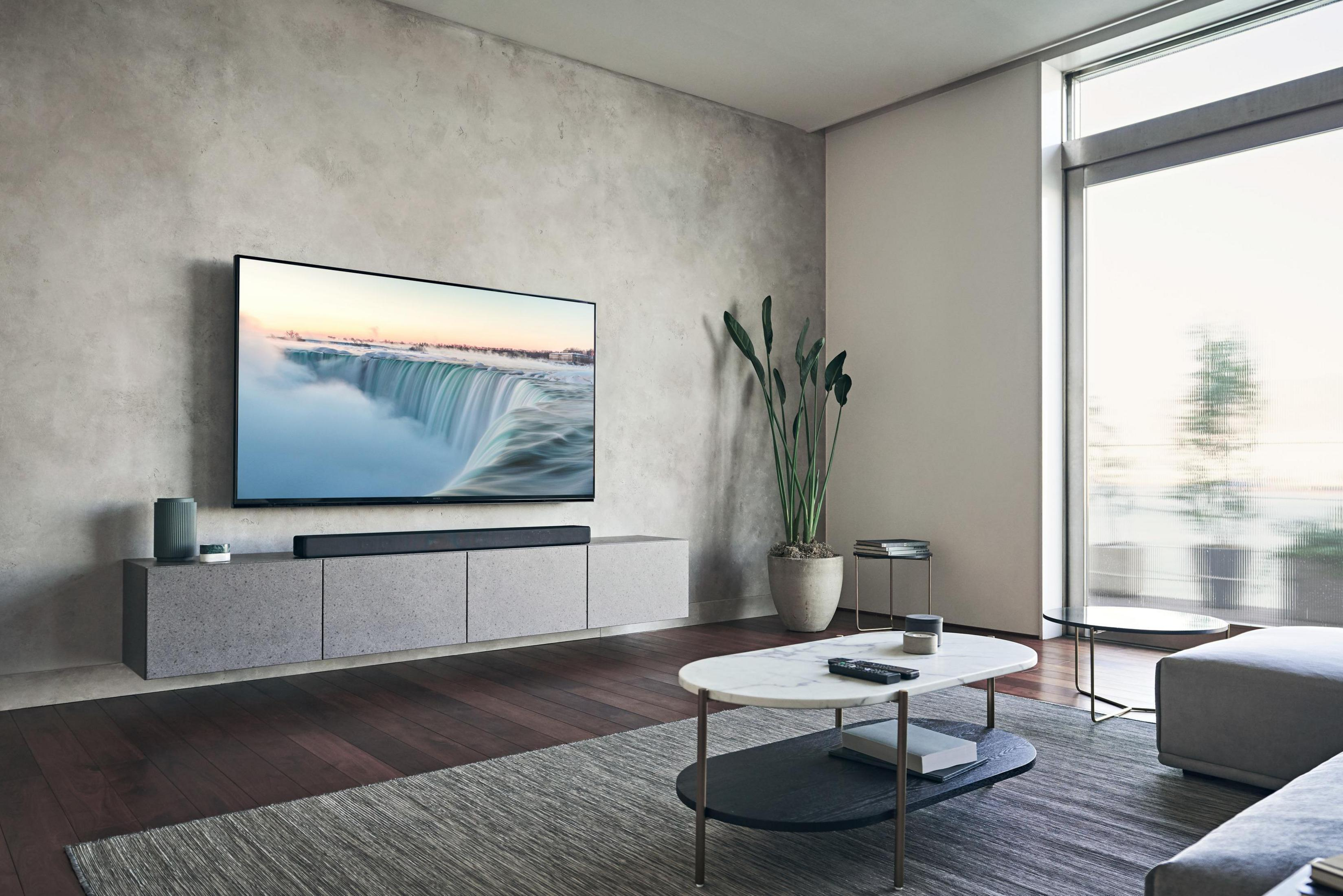 Ein Wohnzimmer mit großem Fernseher und grauen Möbeln.