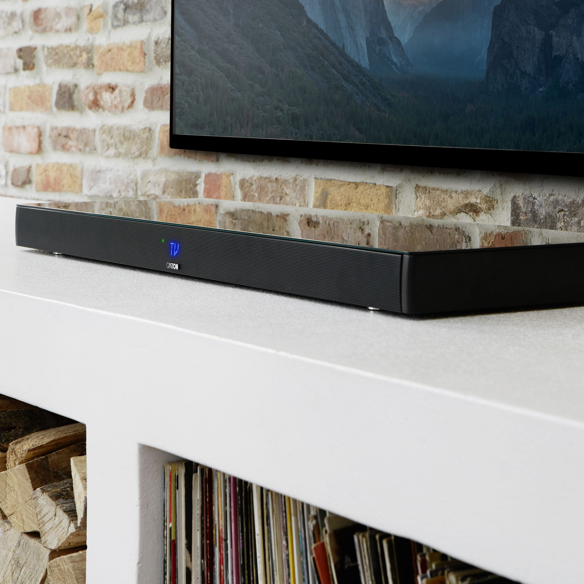 Schwarze Soundbar auf weißem Regal, Ziegelwand im Hintergrund, Fernsehbildschirm, Bücher und Brennholz. Die Soundbar ist sichtbar.