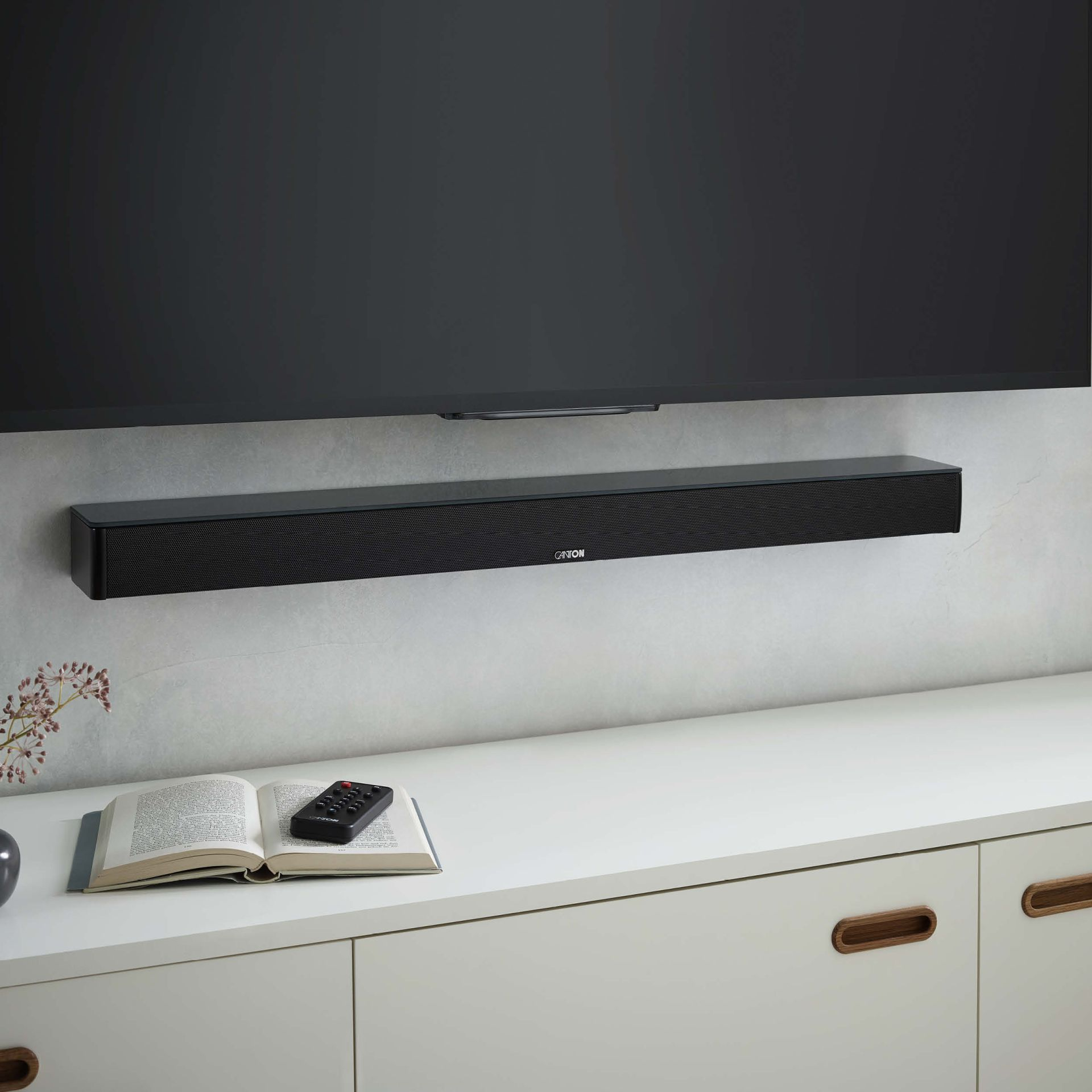 Schwarze Soundbar unter einem Fernseher montiert. Ein Buch und eine Fernbedienung liegen auf dem Schrank.