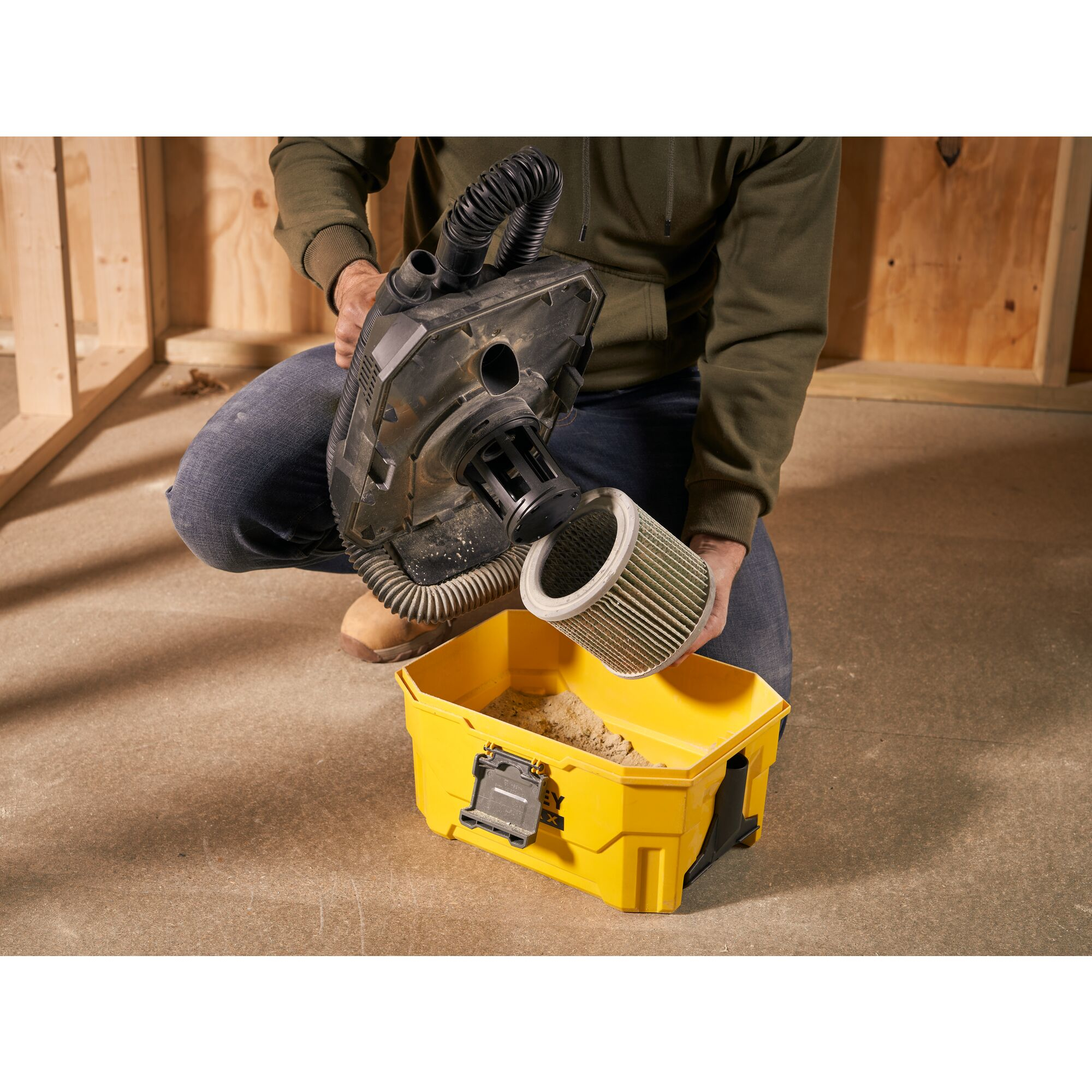 Osoba opróżnia filtr z czarnego odkurzacza do żółtego pojemnika wypełnionego brudem.