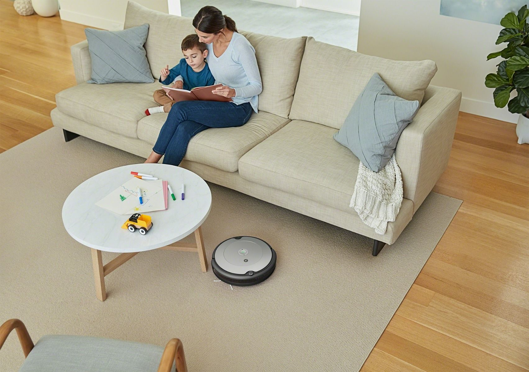 Familie im Wohnzimmer. Mutter und Sohn auf Sofa, lesen. Roboter auf Teppich, Tisch davor.