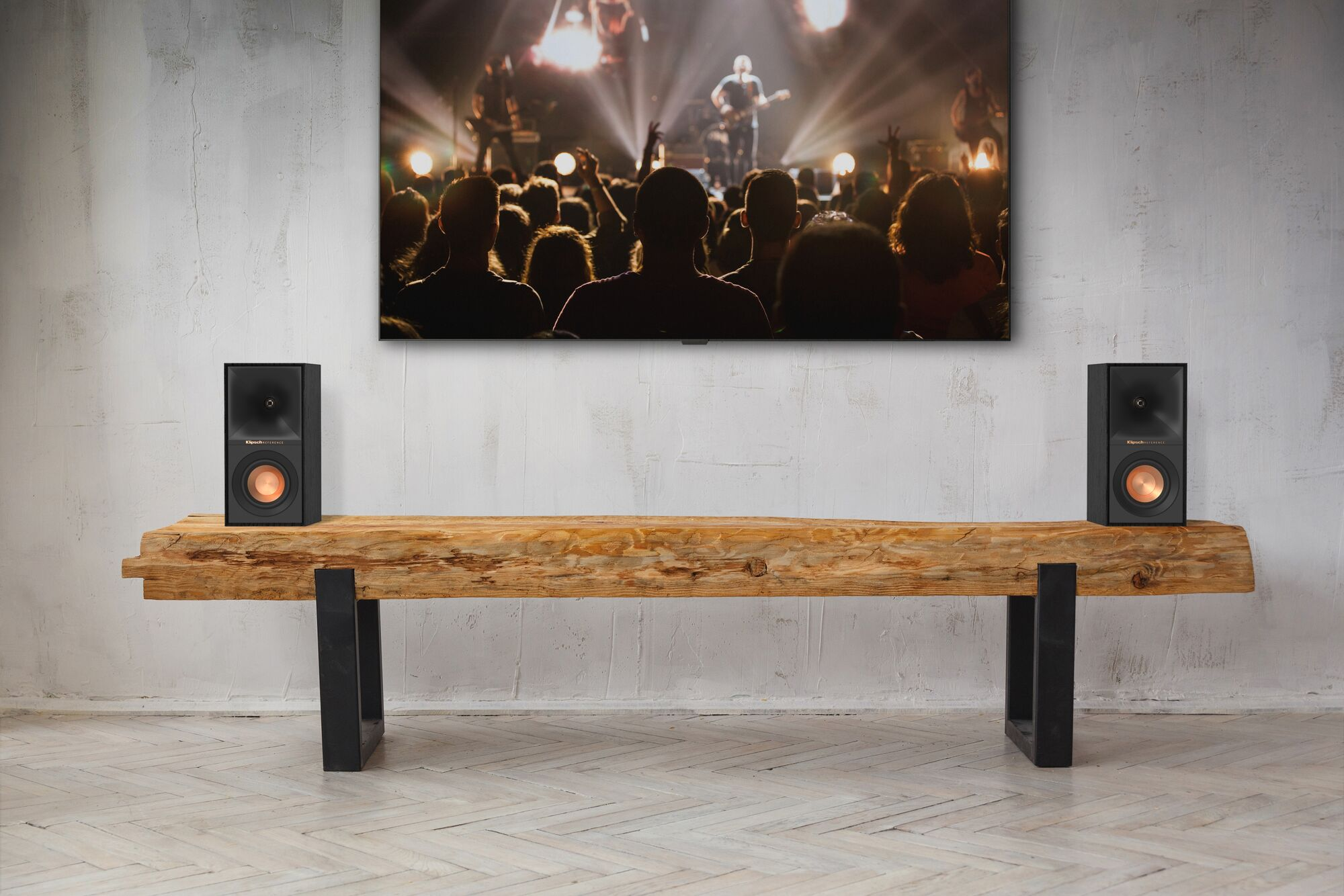 Due altoparlanti su un tavolo di legno sotto una TV che mostra un concerto. Lo sfondo è un muro.