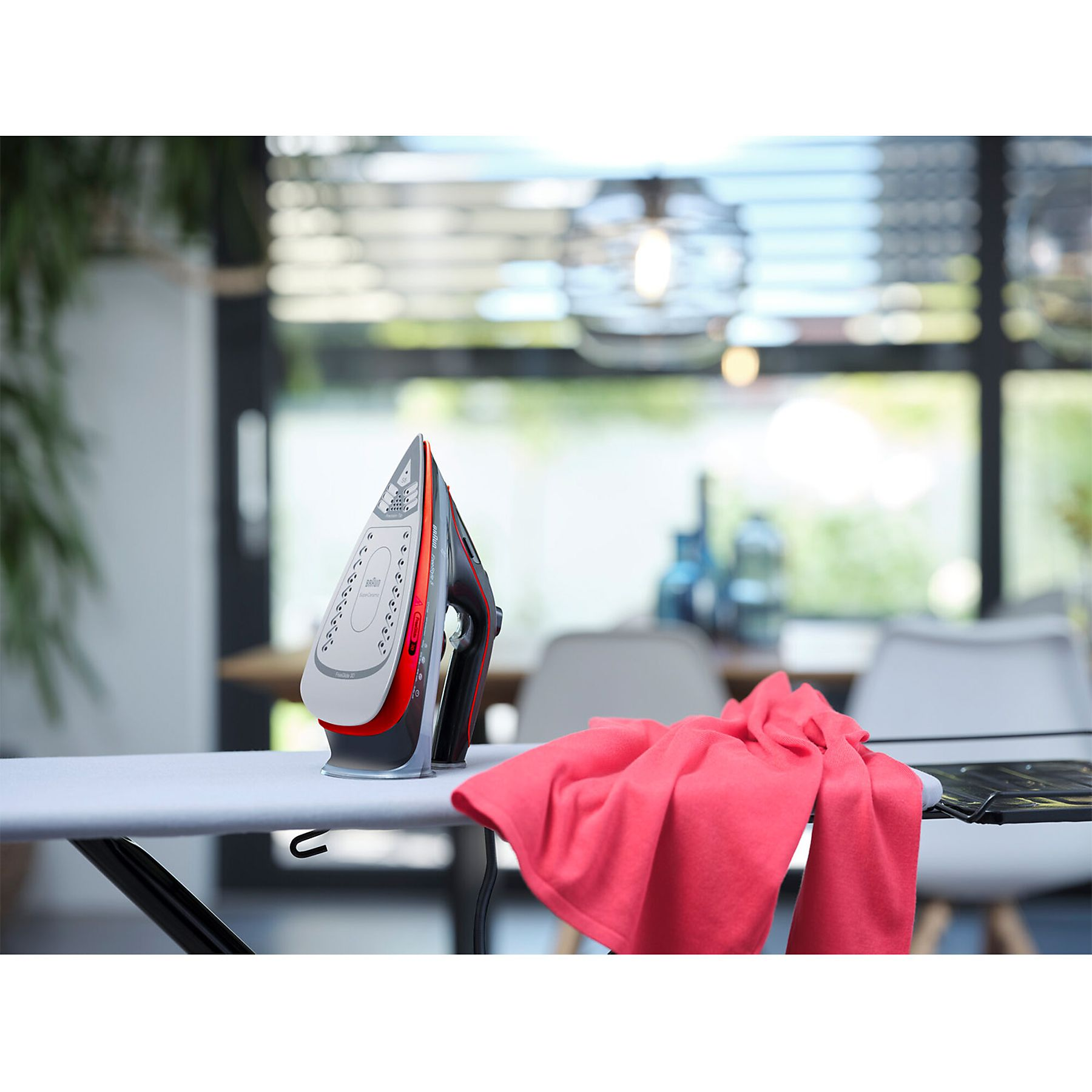 Deska do prasowania z żelazkiem i czerwonymi ubraniami. Stół i okno są w tle.