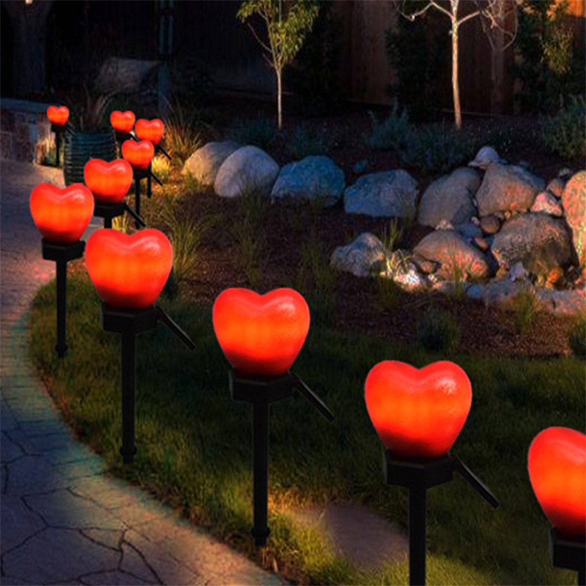 Rote herzförmige Lichter auf schwarzen Pfählen in einem Garten. Grünes Gras und Pflanzen im Hintergrund.