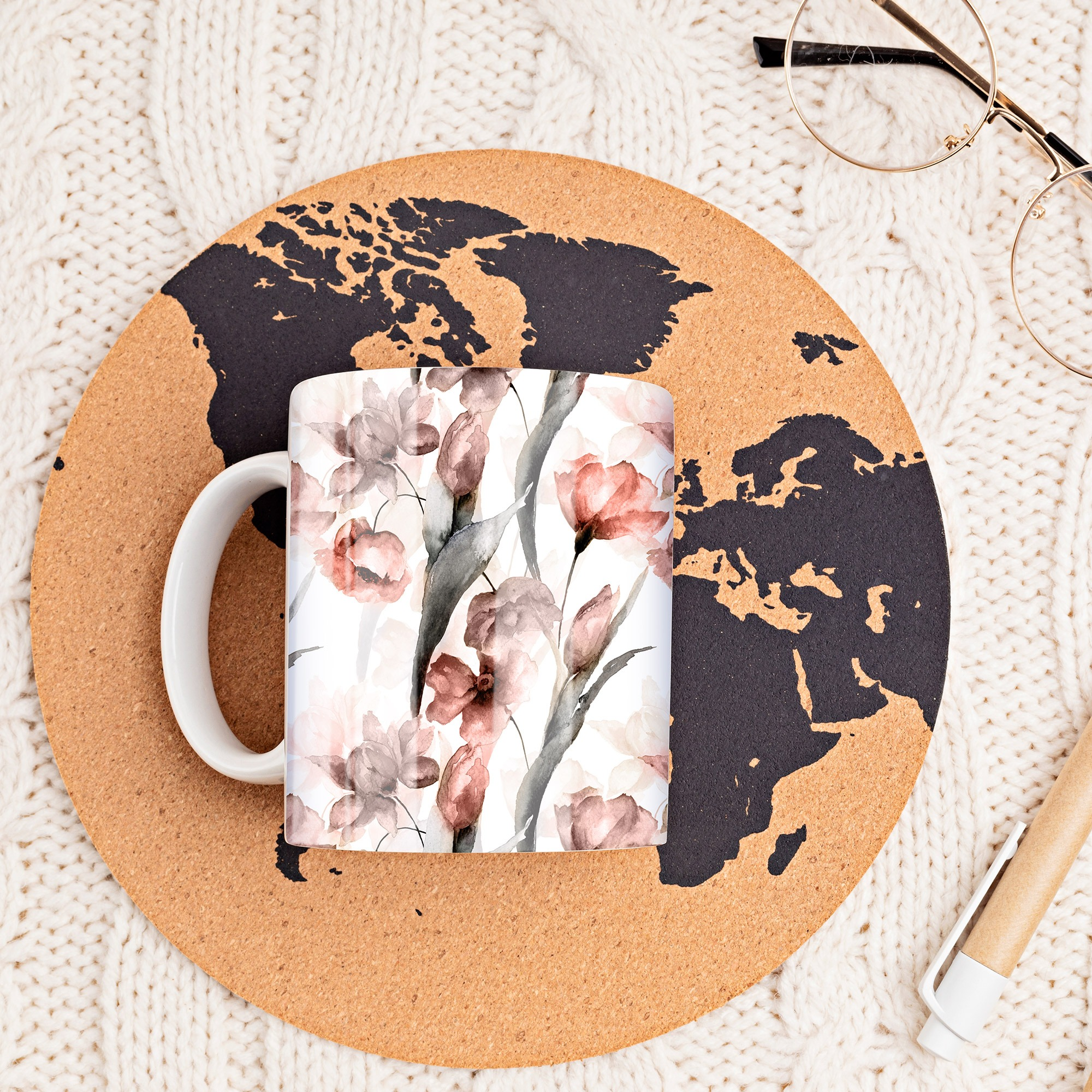 Becher mit floralem Design auf Korkuntersetzer über weißer Strickware, mit Brille und Stift.