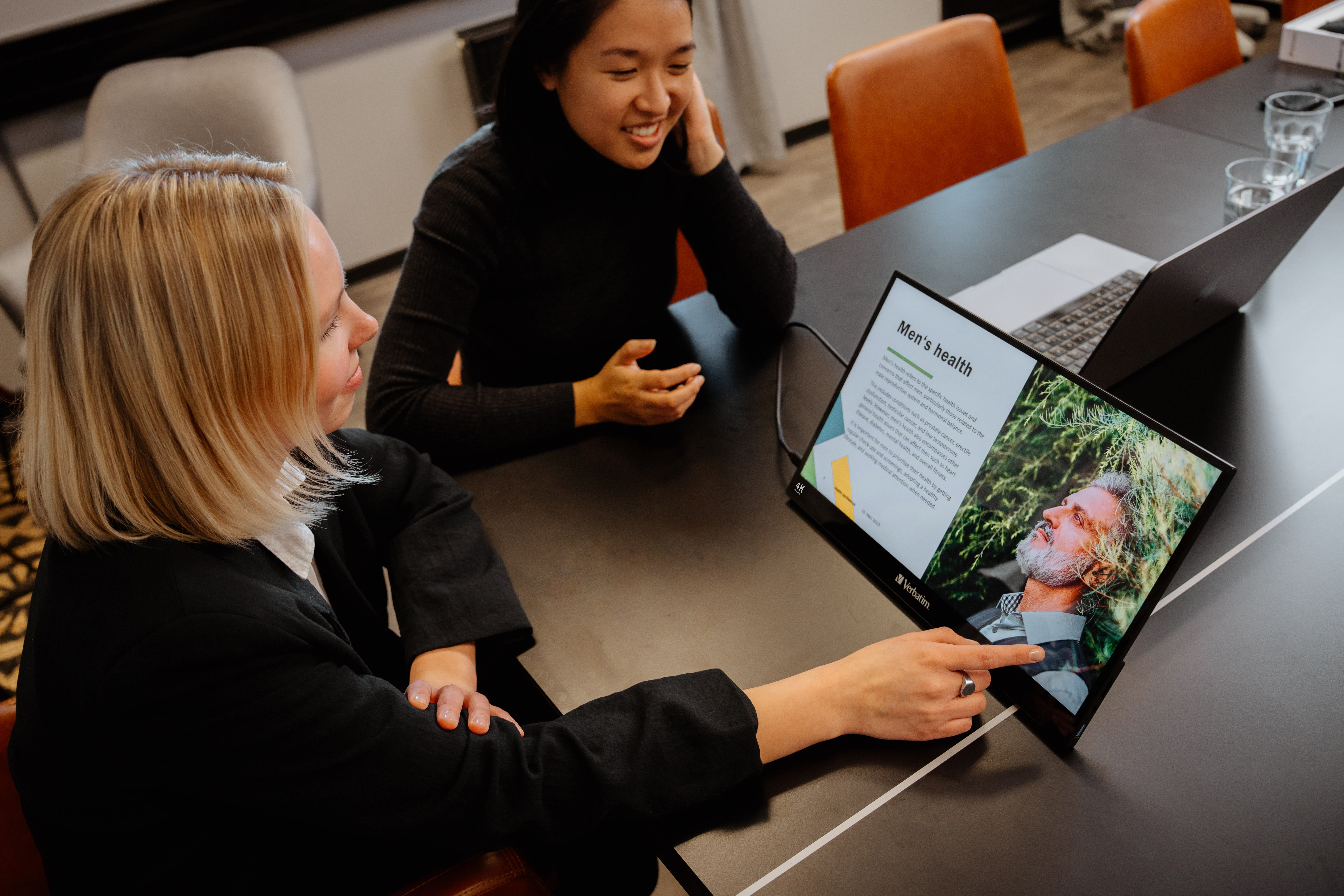 Dwie kobiety siedzą przy stole. Jedna wskazuje na ekran z prezentacją na temat zdrowia mężczyzn.