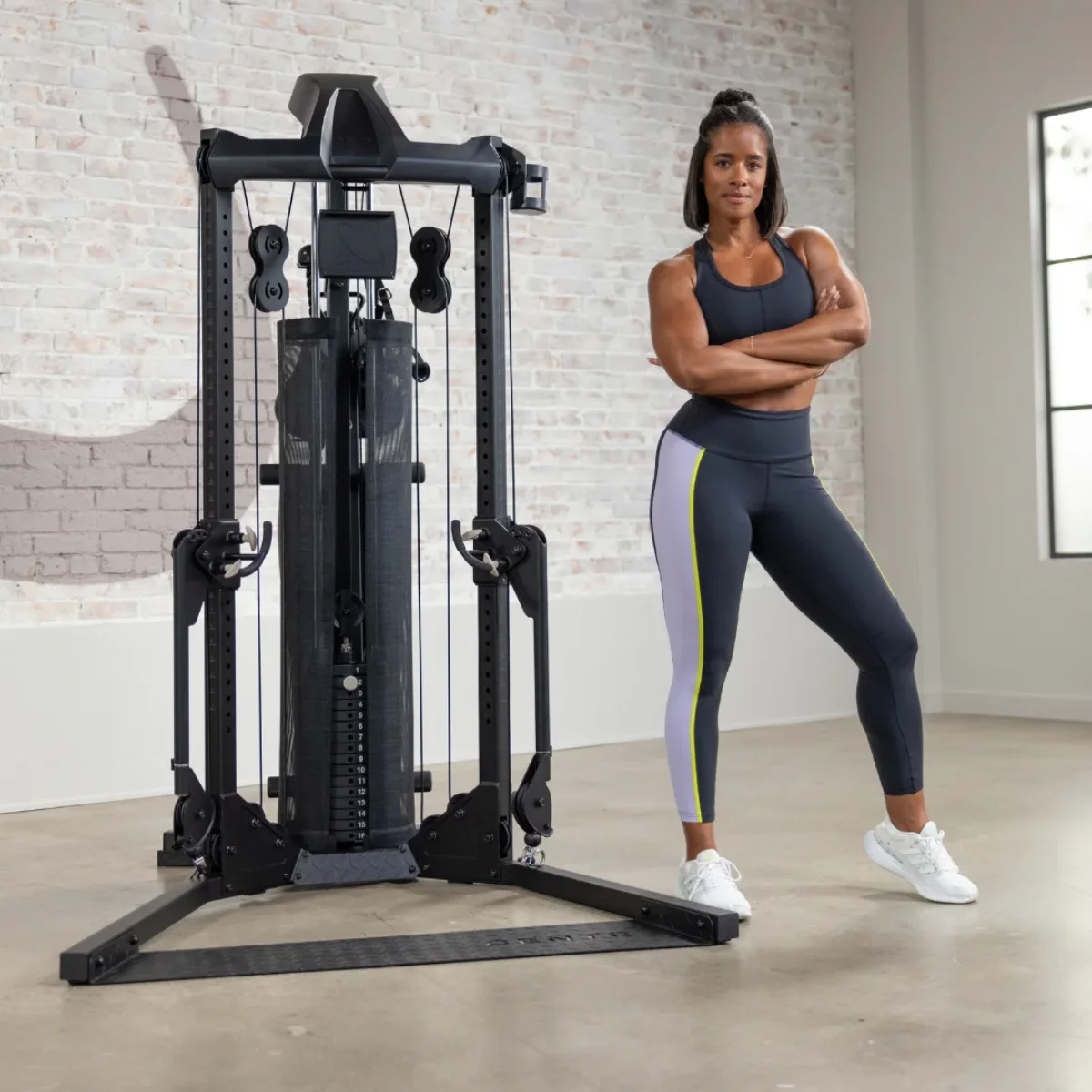 Frau in Sportkleidung steht neben einer schwarzen Fitnessmaschine. Ziegelwand im Hintergrund.