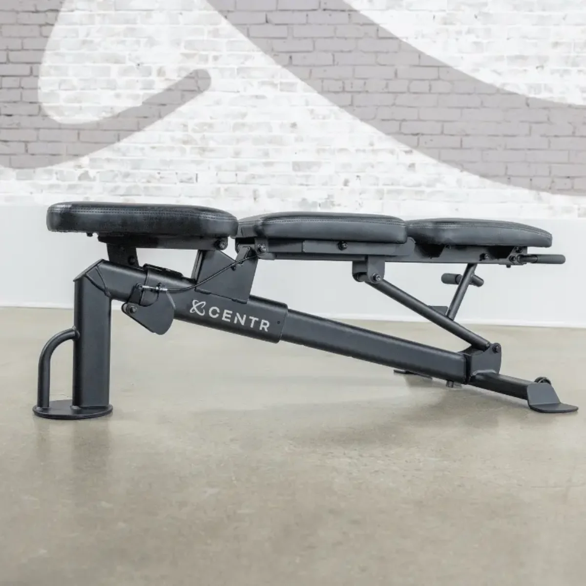 A black weight bench with adjustable angles, set against a white brick wall.