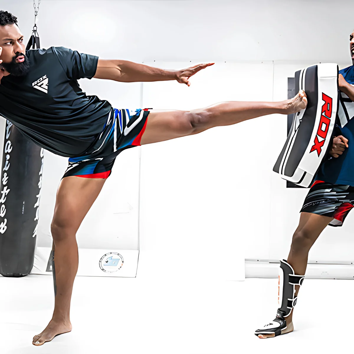 Two men in a gym. One is kicking a pad held by the other. Both are bare-footed.