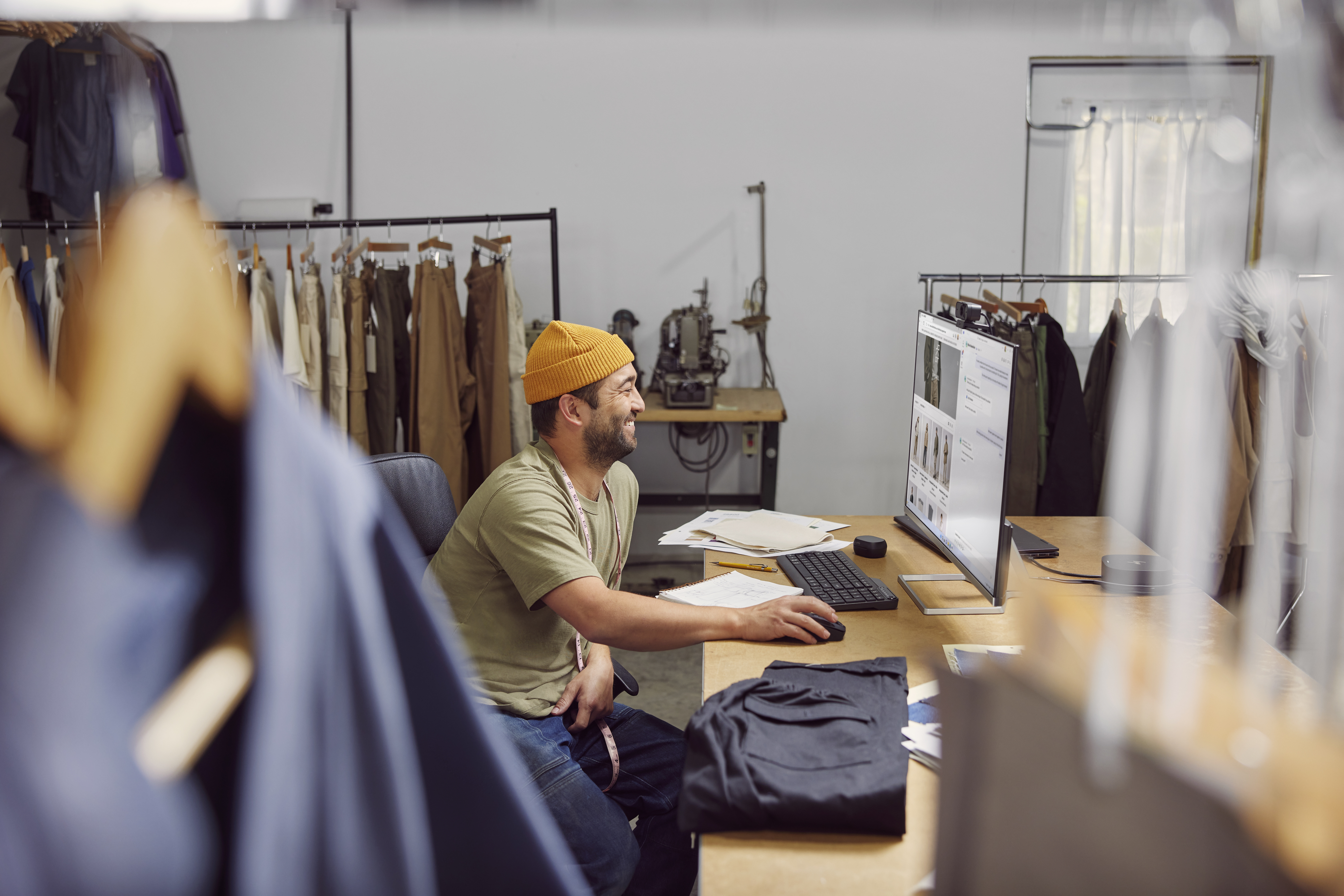 Mężczyzna w warsztacie z komputerem, żółty kapelusz, uśmiechnięty. Ubrania na wieszakach, maszyna do szycia, biurko z papierami, światło.