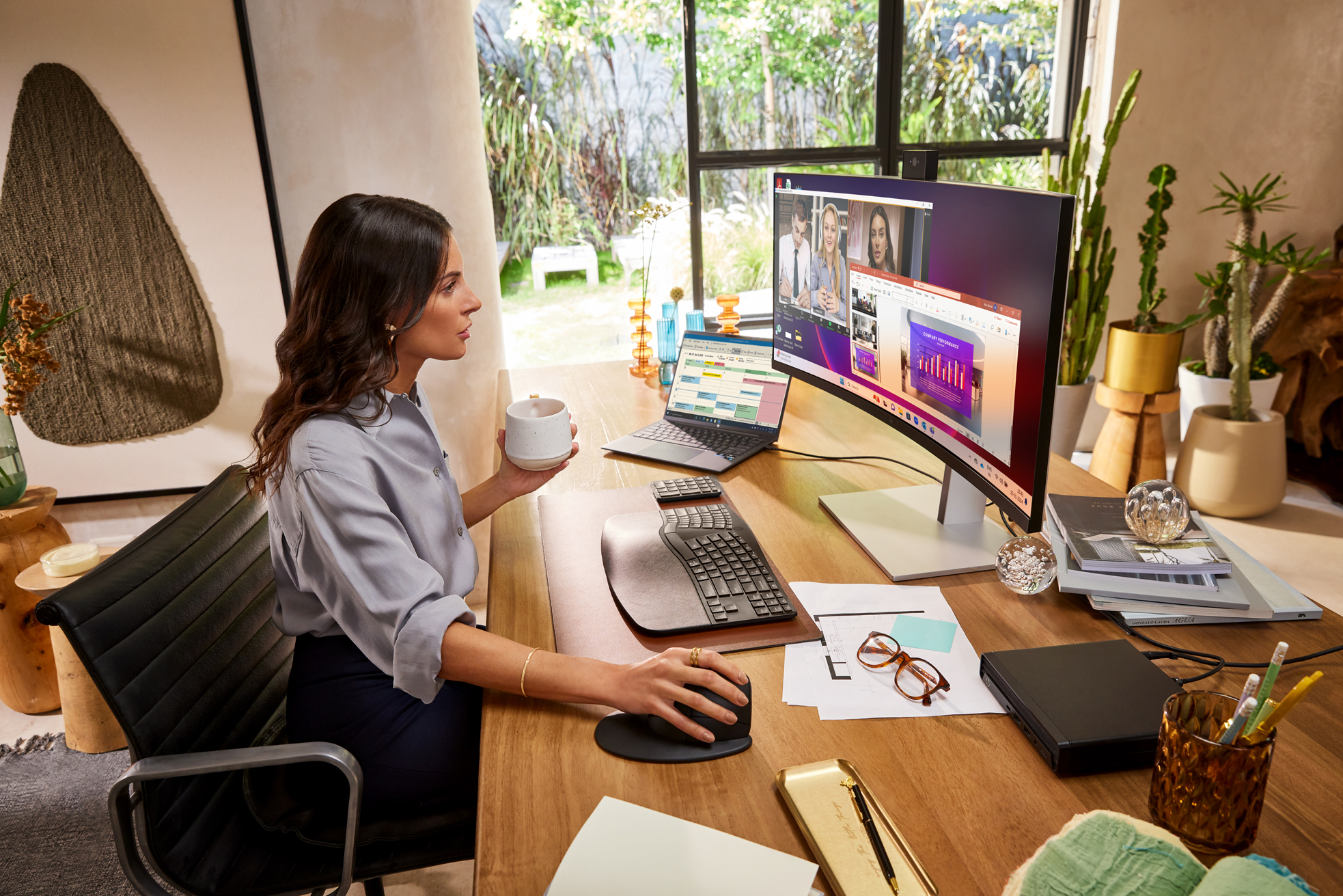 Kobieta przy biurku z dwoma monitorami, laptopem i kubkiem. Jest na wideorozmowie.