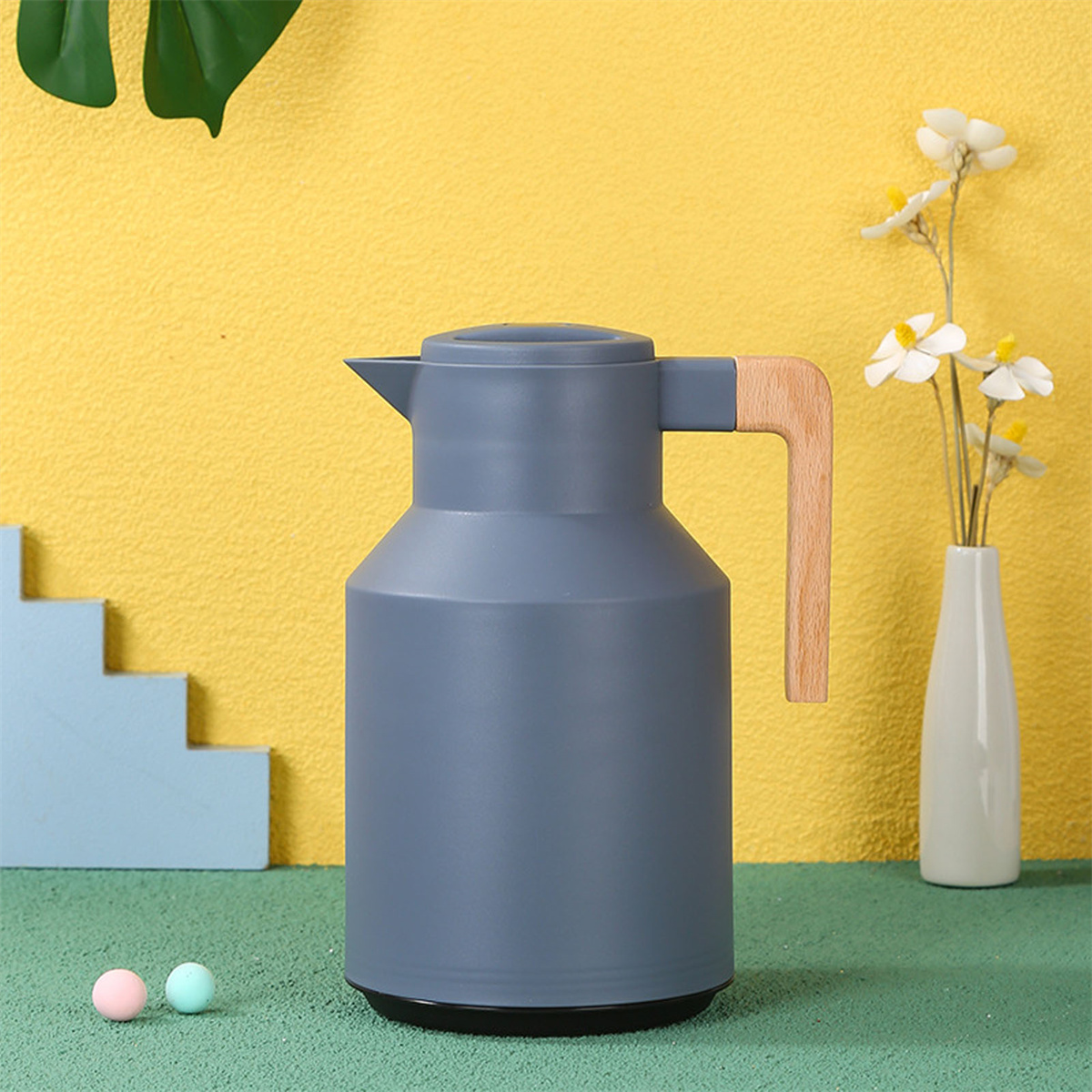 Eine graue Thermoskanne mit Holzgriff und eine Vase mit Blumen, auf einer grünen Oberfläche mit pinken und blauen Kugeln.