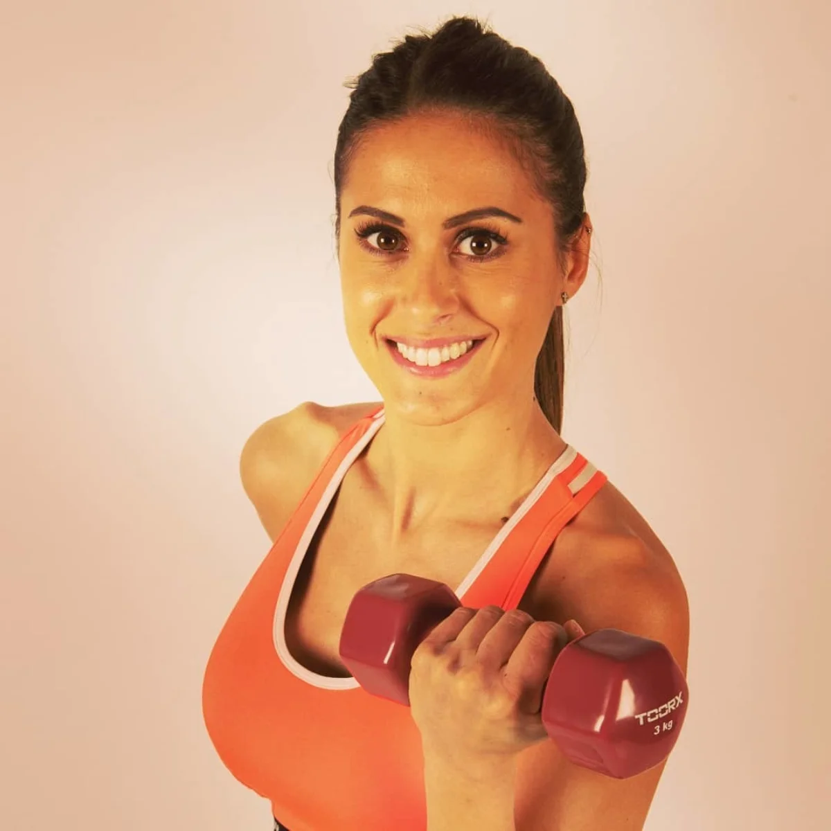 Eine lächelnde Frau hält eine weinrote Kurzhantel, orange Sport-BH, weißer Hintergrund.