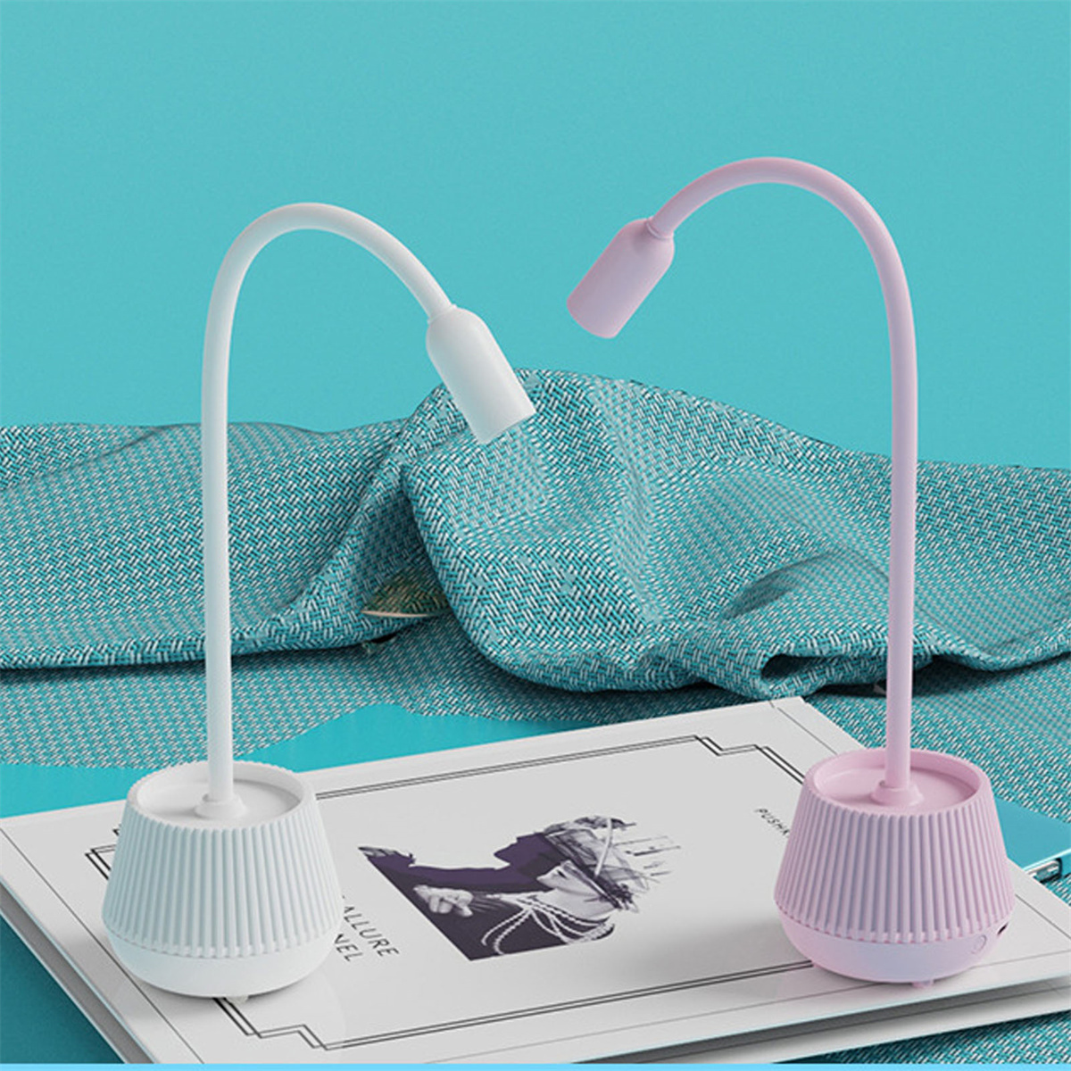 Zwei kleine Schreibtischlampen stehen auf einem Notizbuch. Eine ist weiß, die andere rosa, beide mit gebogenen Hälsen.