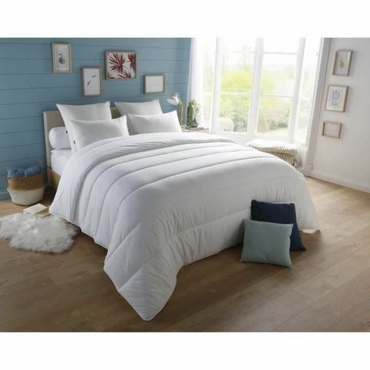 Ein Schlafzimmer mit Bett, Kissen und weißer Bettdecke. Eine blaue Wand und ein Fenster mit Sonnenlicht sind sichtbar.