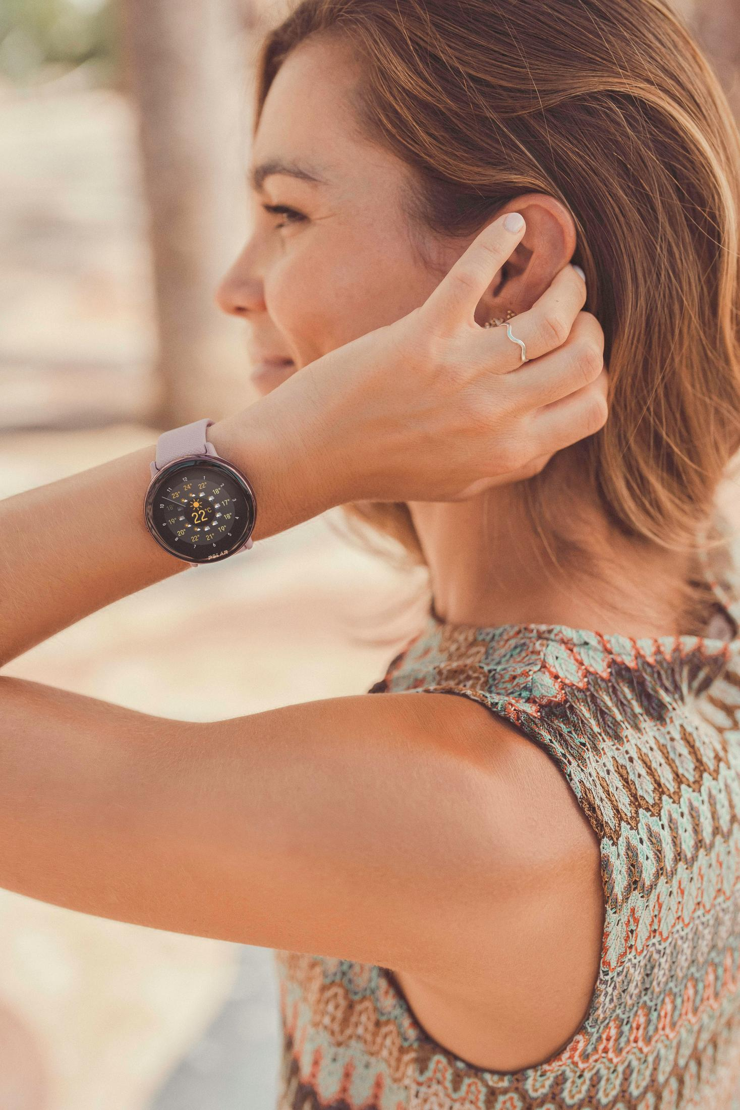 Kobieta nosząca różowy smartwatch, dotykająca ucha, z rozmytym tłem.