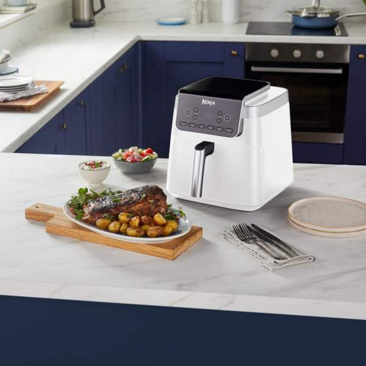 Frytownica powietrzna na blacie kuchennym z ugotowanym jedzeniem na drewnianym talerzu. Niebieskie szafki w tle.