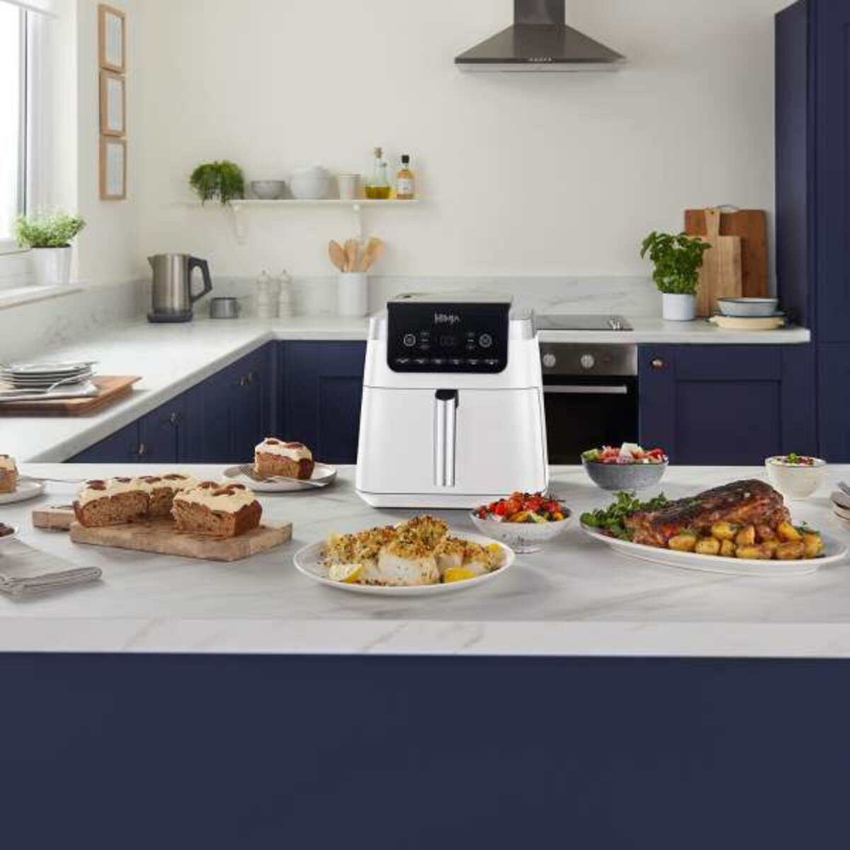 Kuchnia z frytkownicą powietrzną na blacie, różne potrawy i ciemnoniebieskie szafki.